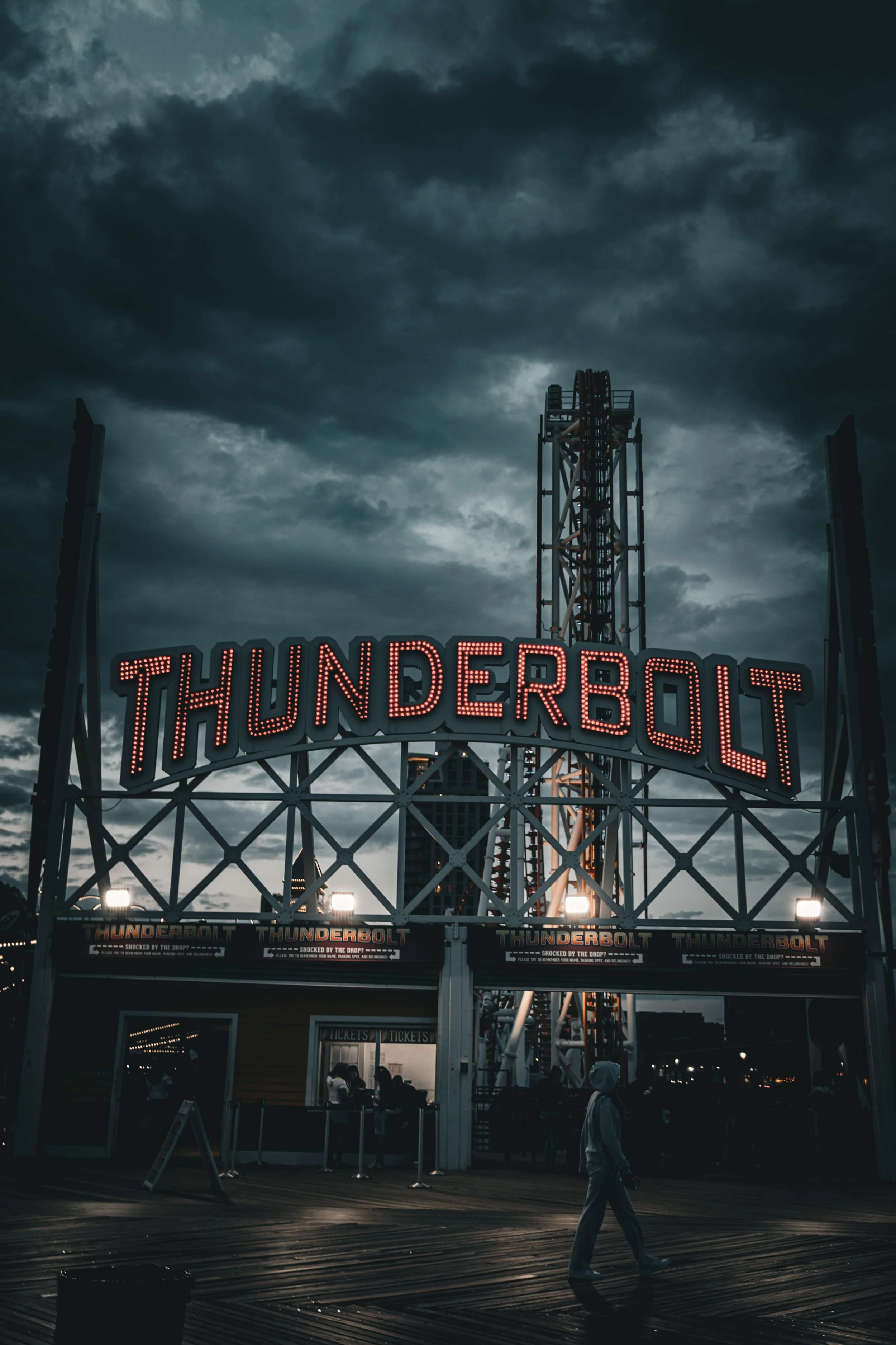 A dark sky with storm clouds over a carnival ride named Thunderbolt, illuminated by red lights and lights at the bottom, with a person walking in the foreground.