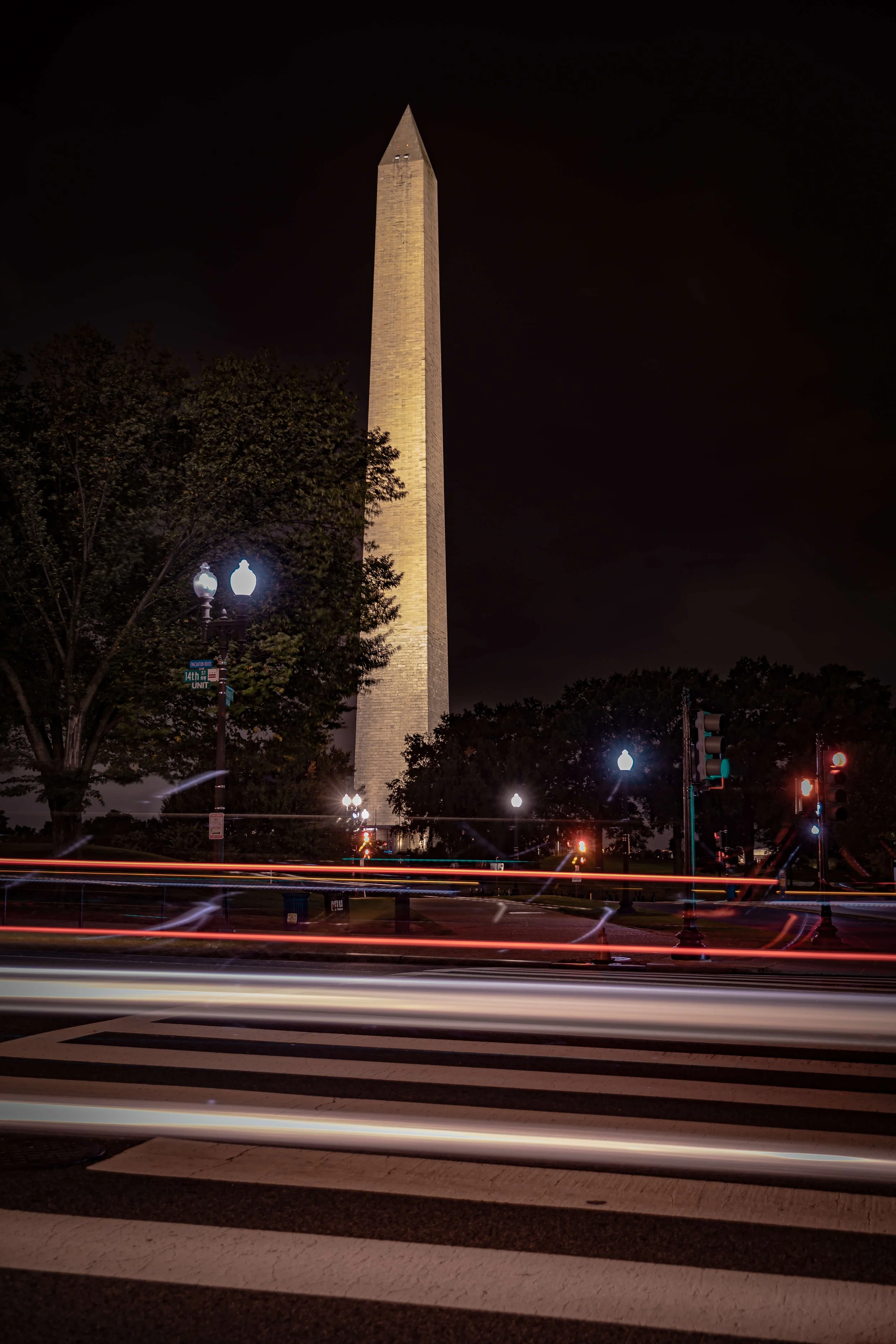 Nighttime view of the Washington Monument in Washington, D.C., with light trails from passing cars on a crosswalk in the foreground.