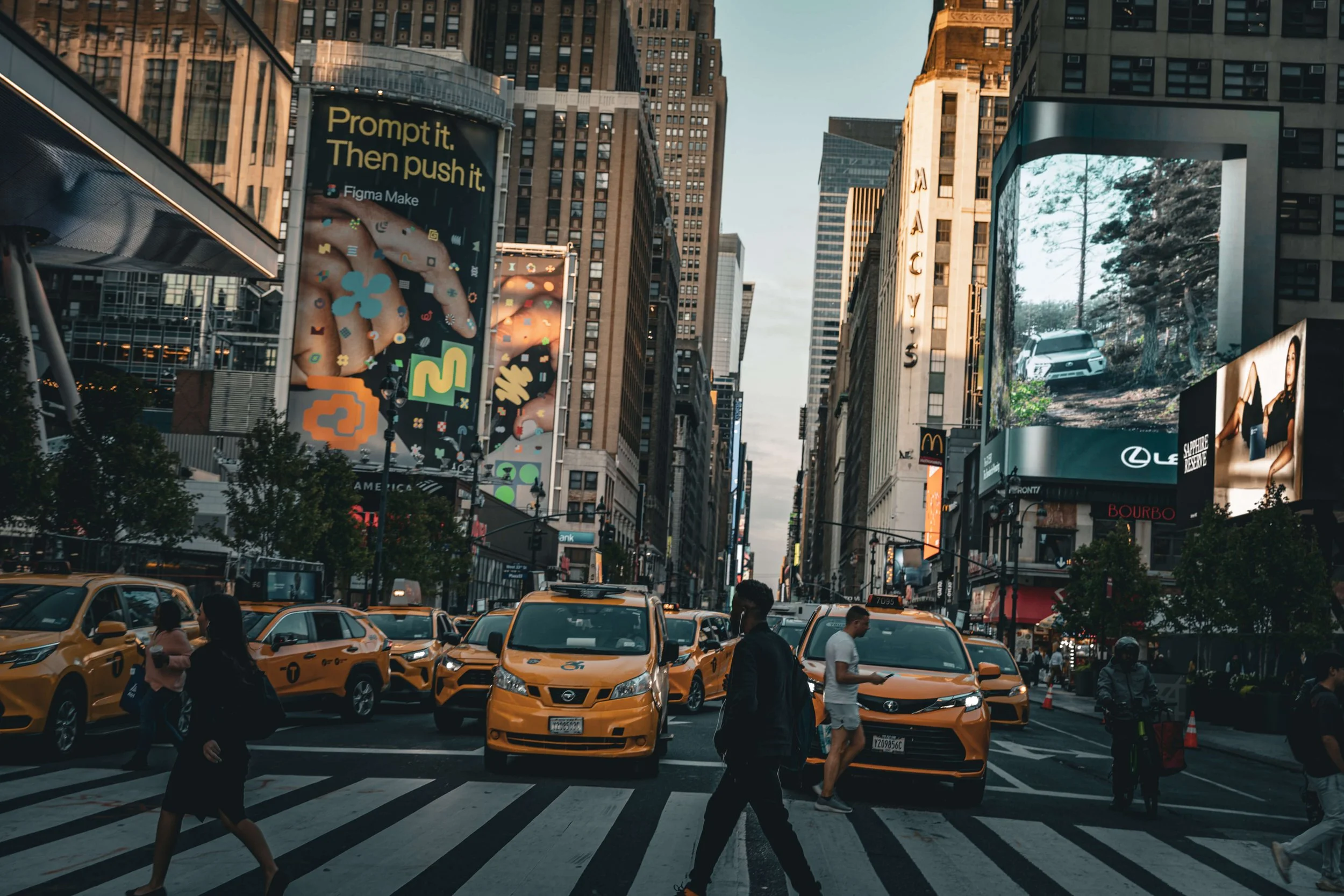 City street scene in New York City with yellow taxis, pedestrians crossing at a crosswalk, tall buildings with billboards, and advertisements, including one for McDonald's and Lexus.