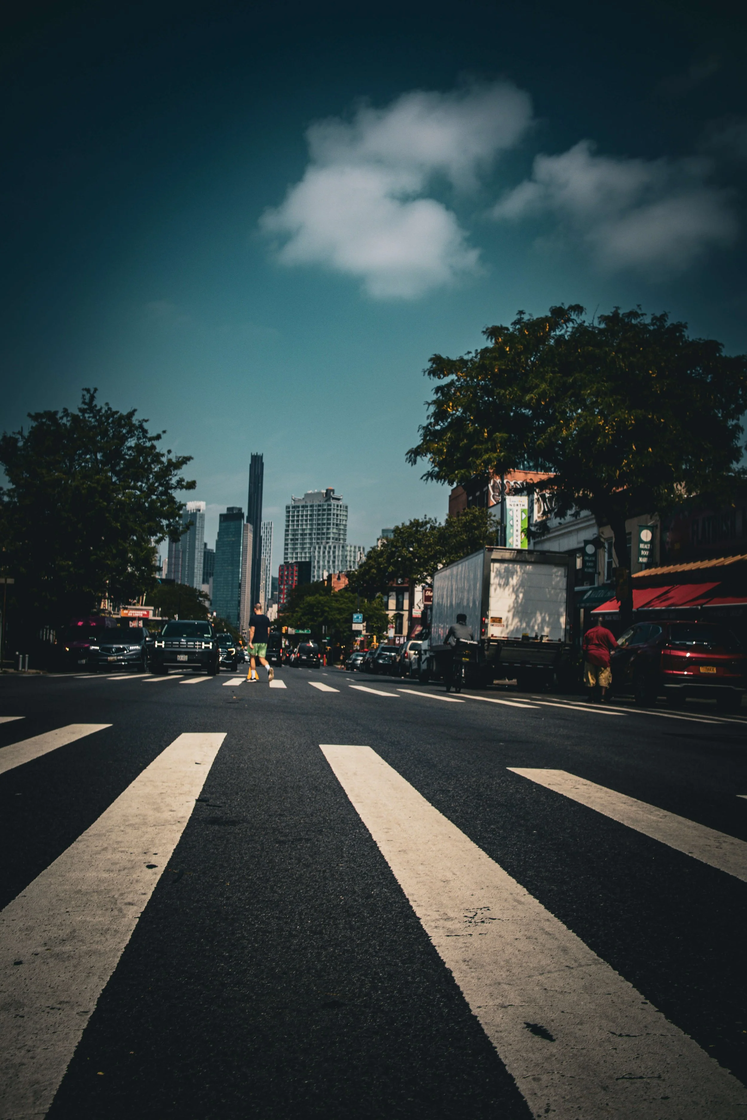 City street view with crosswalk, pedestrians, parked cars, trees lining the street, and tall skyscrapers in the background under a partly cloudy sky.