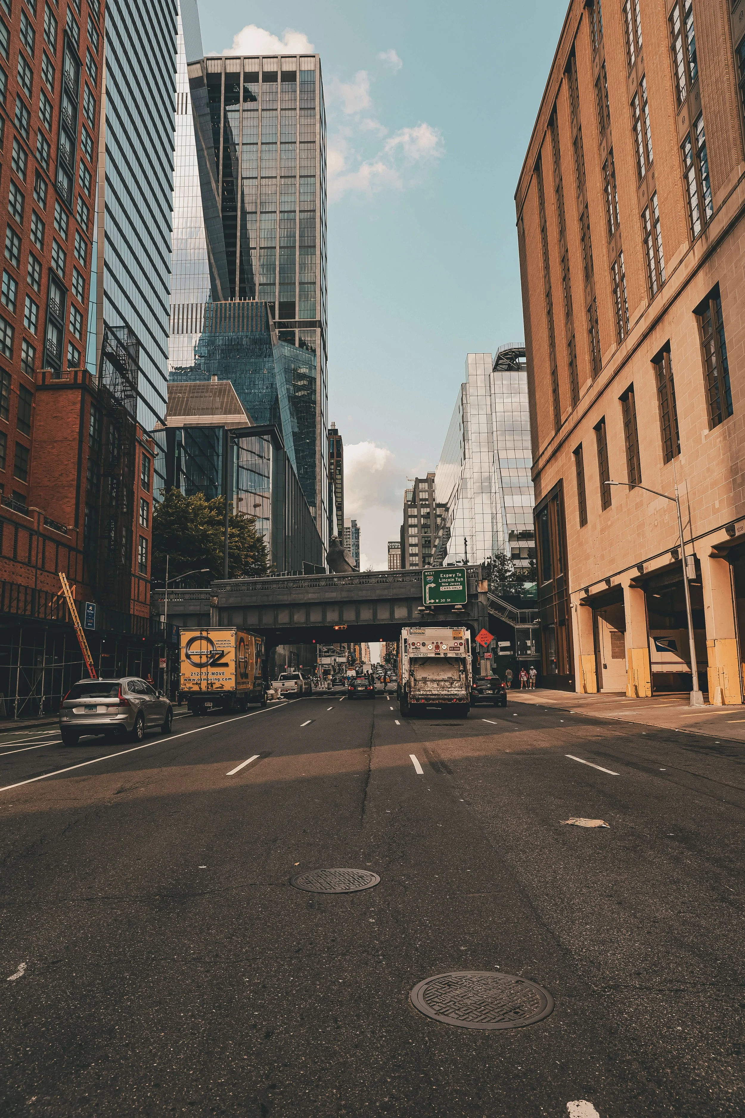City street scene with tall skyscrapers on both sides, cars on the road, and a footbridge crossing above the street in a downtown area.