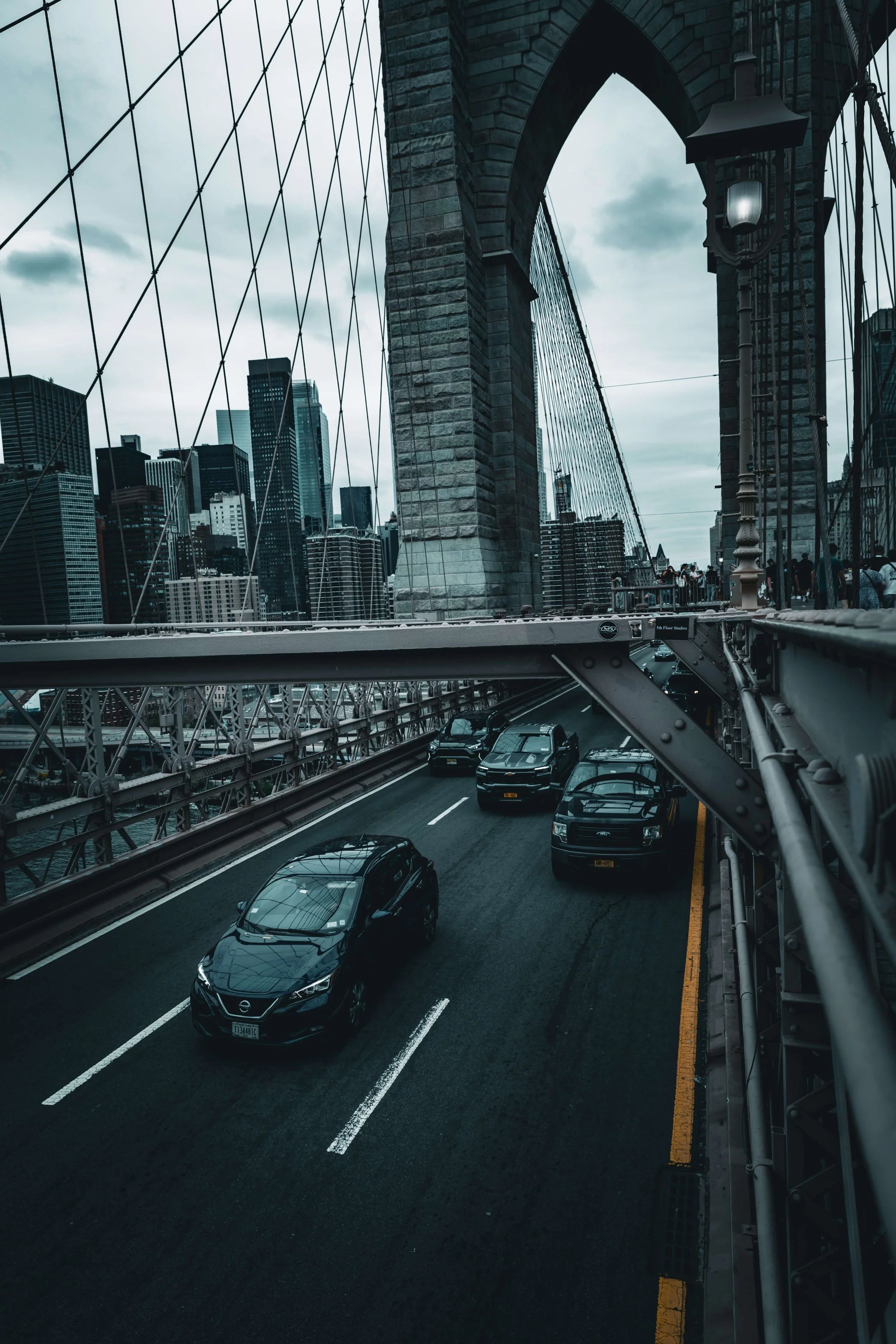 View of a city bridge with cars driving on the road beneath the bridge structure, high-rise buildings in the background, and cloudy sky overhead.
