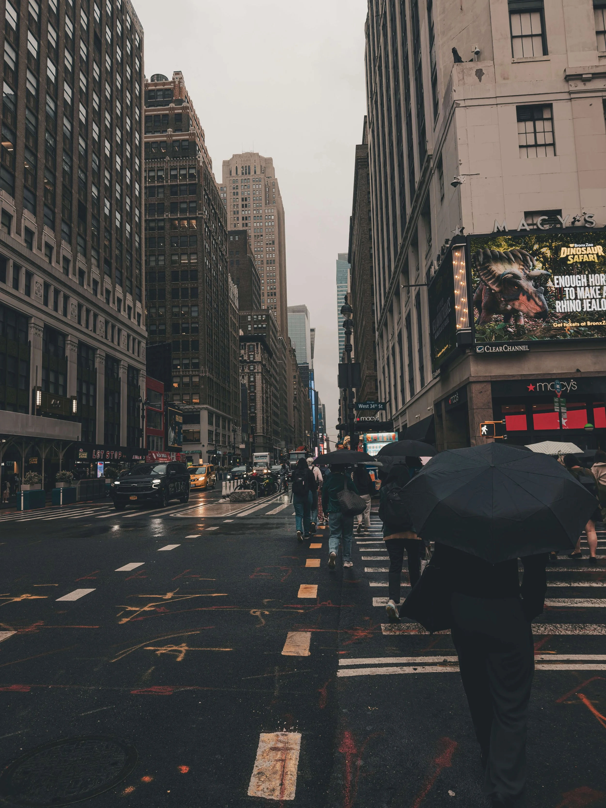 People walking with umbrellas on a city street during rainy weather, surrounded by tall buildings and electronic billboards.