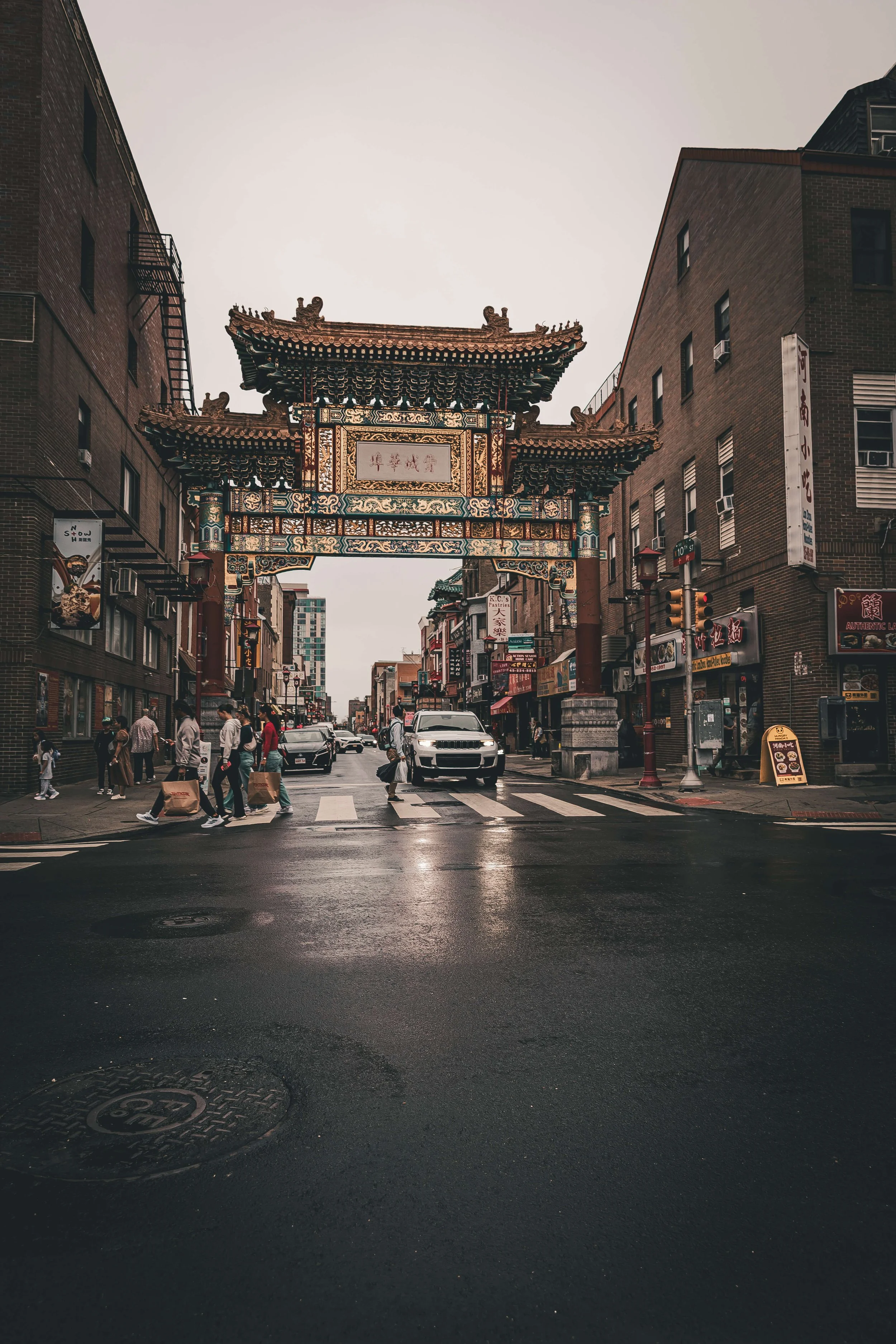 View of a city street with a traditional Chinese gate archway at the intersection, surrounded by brick buildings and pedestrians crossing the street on a rainy day.