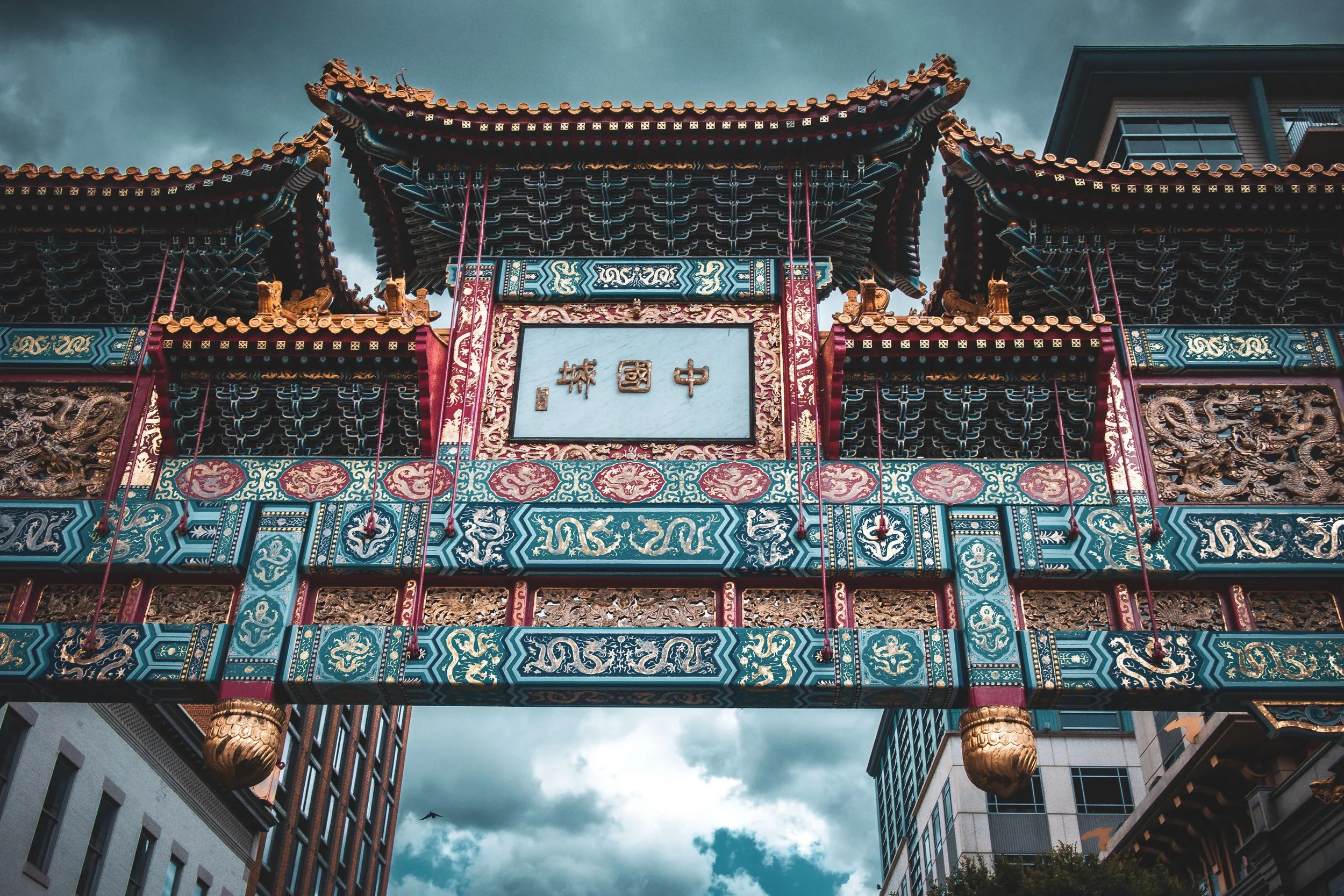 A traditional Chinese decorative gateway with colorful intricate designs, dragons, and gold accents, set against a cloudy sky with modern buildings on either side.