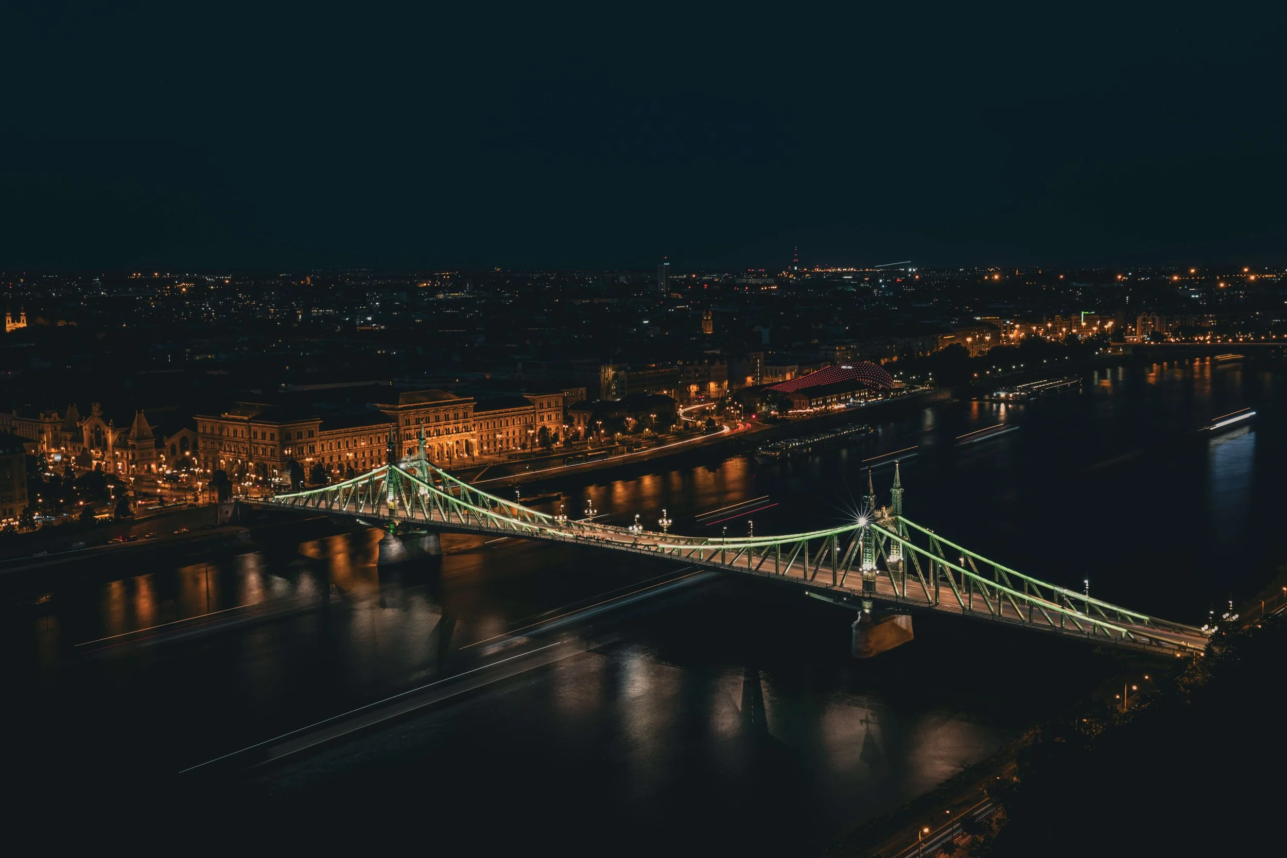 Nighttime cityscape with illuminated bridge crossing a river, historic buildings along the waterfront, and a city skyline in the background.