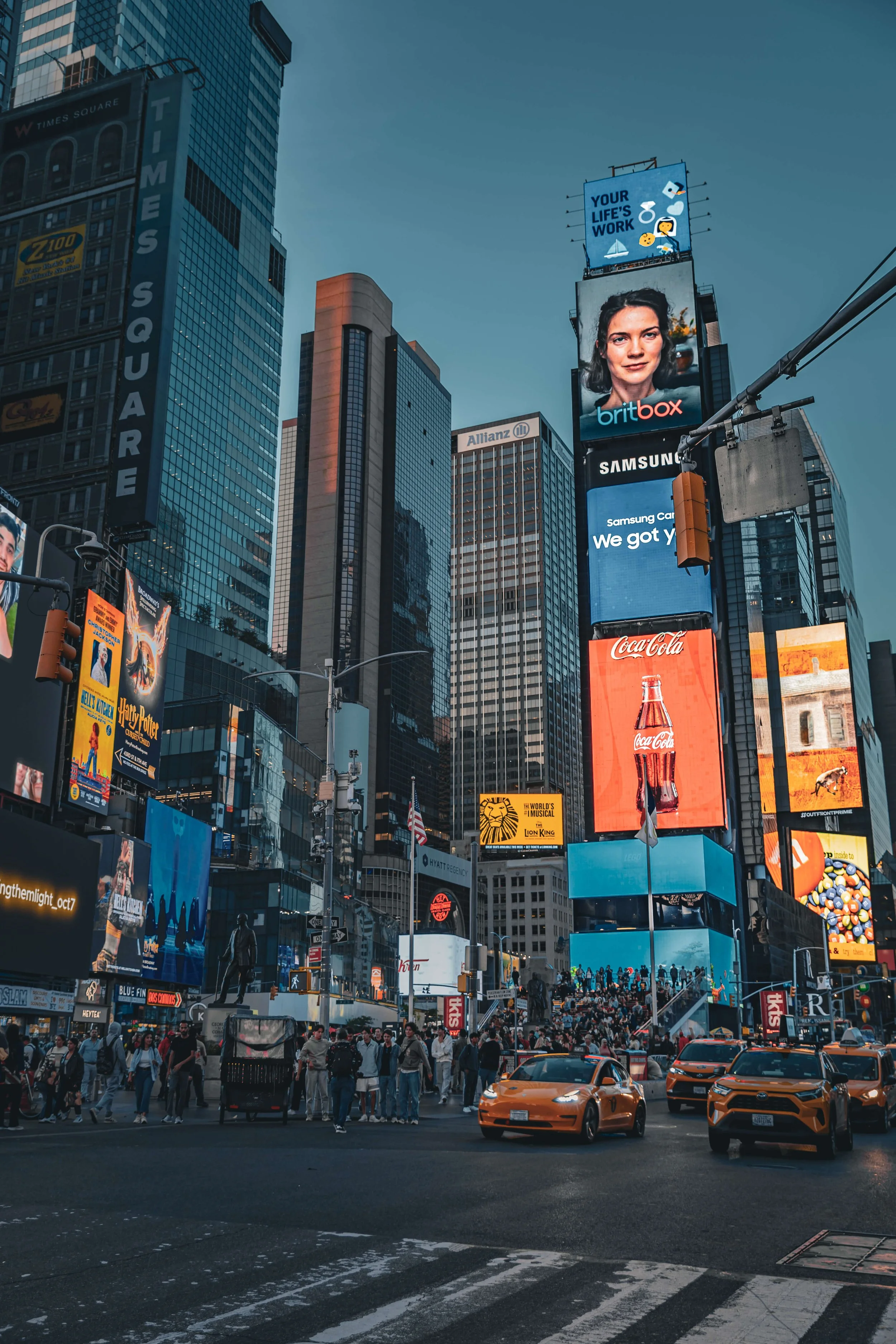 Times Square in New York City at dusk, featuring tall buildings with bright electronic billboards, yellow taxis on the street, and a crowd of pedestrians.