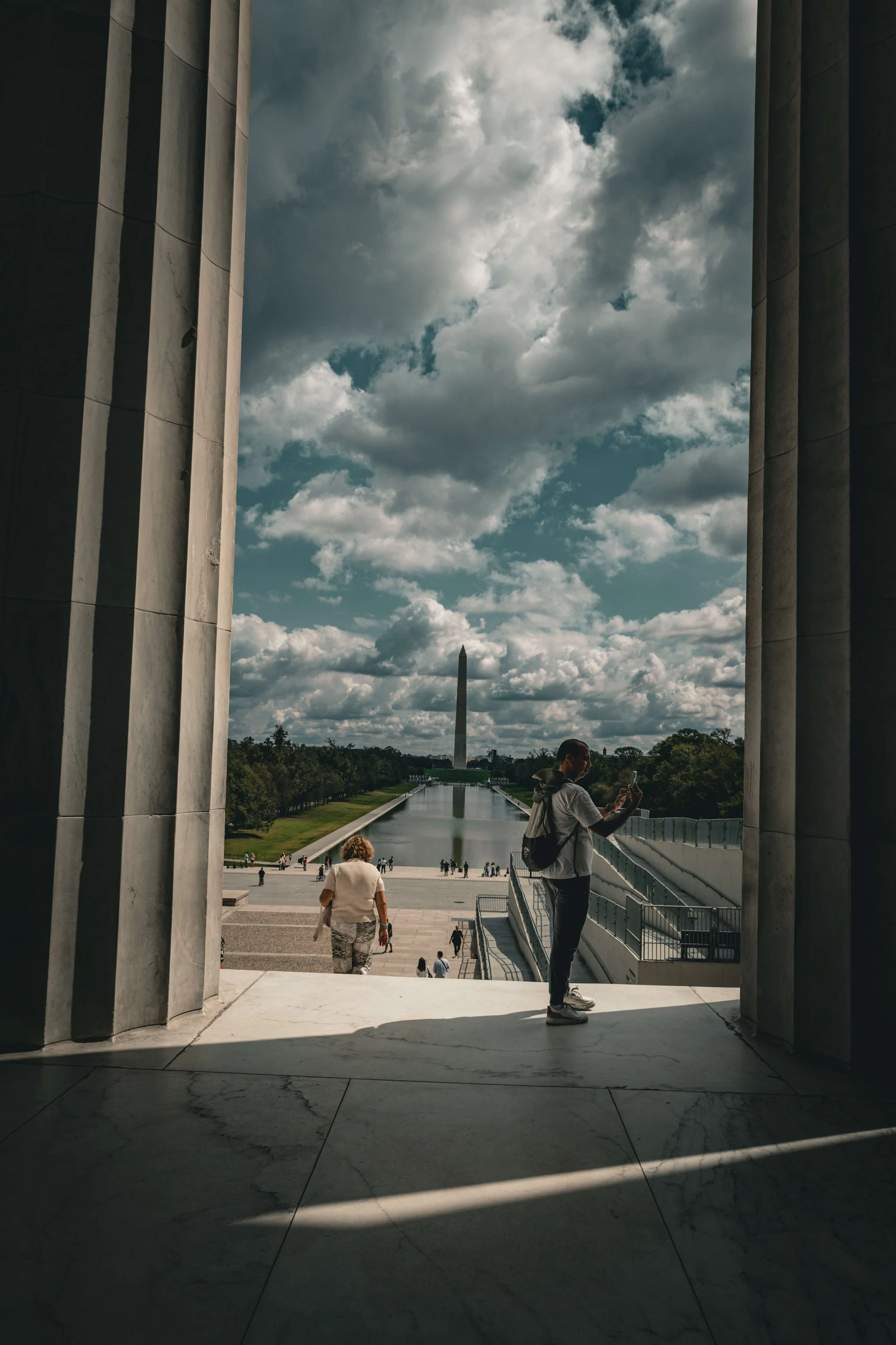 View of the Washington Monument through the arch of the Lincoln Memorial in Washington, D.C. with people taking photos and walking near the reflecting pool under a partly cloudy sky.