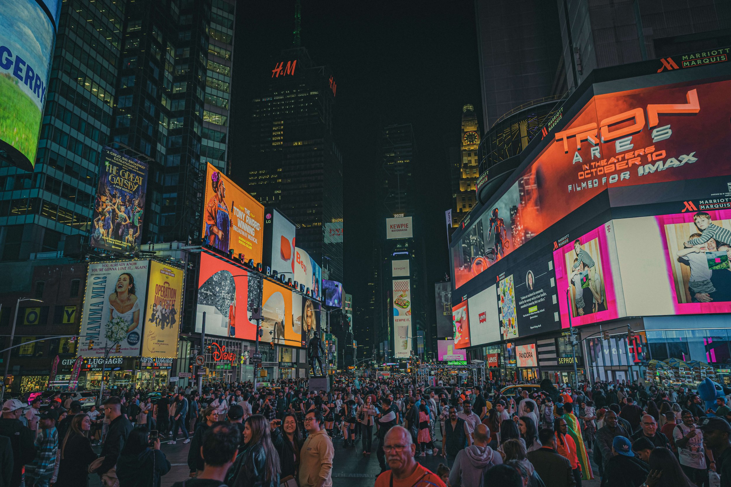A crowded city square at night with illuminated billboards and skyscrapers, including advertisements for theater, fashion, and entertainment.