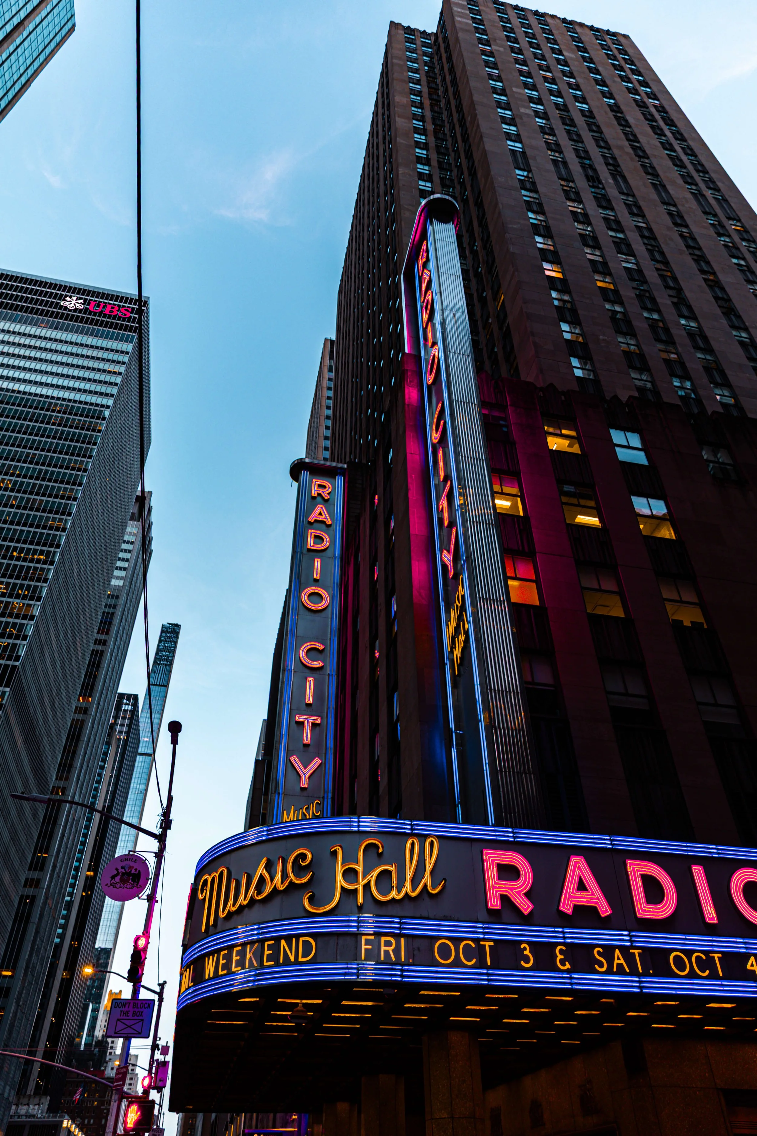 The image shows the marquee of a Broadway theater decorated with colorful neon lights, advertising 'Music Hall' and 'Radio City' in Times Square, New York City, during twilight.