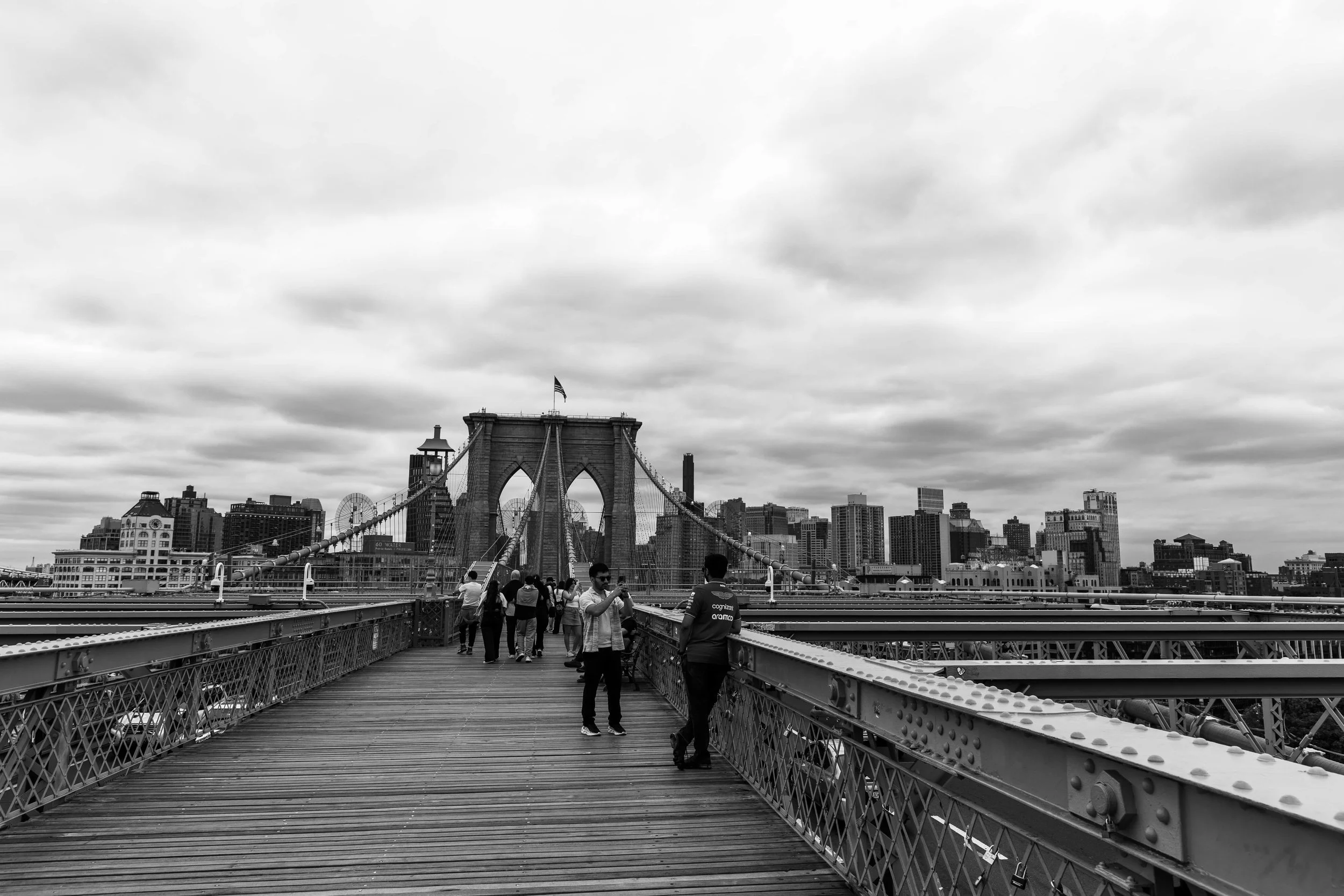 Black and white photo of the Brooklyn Bridge with people walking on the pedestrian walkway and New York City skyline in the background.