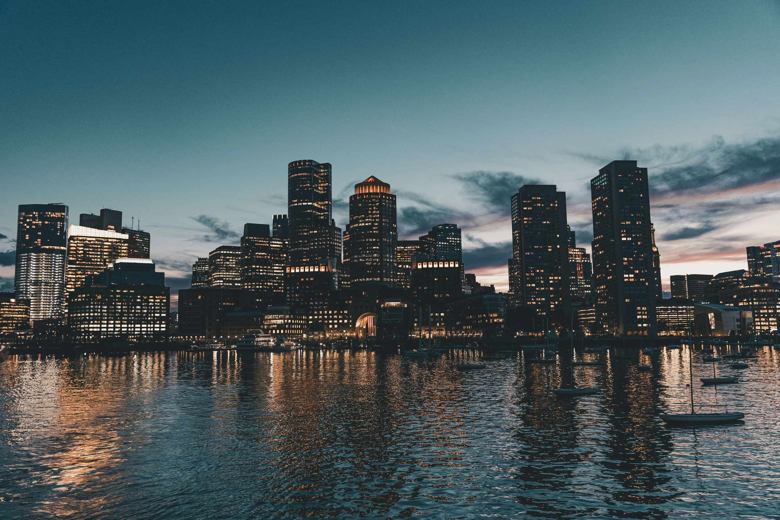 City skyline at dusk with tall office buildings, some illuminated, along the waterfront with small sailboats on the water, and a sky with dark clouds.