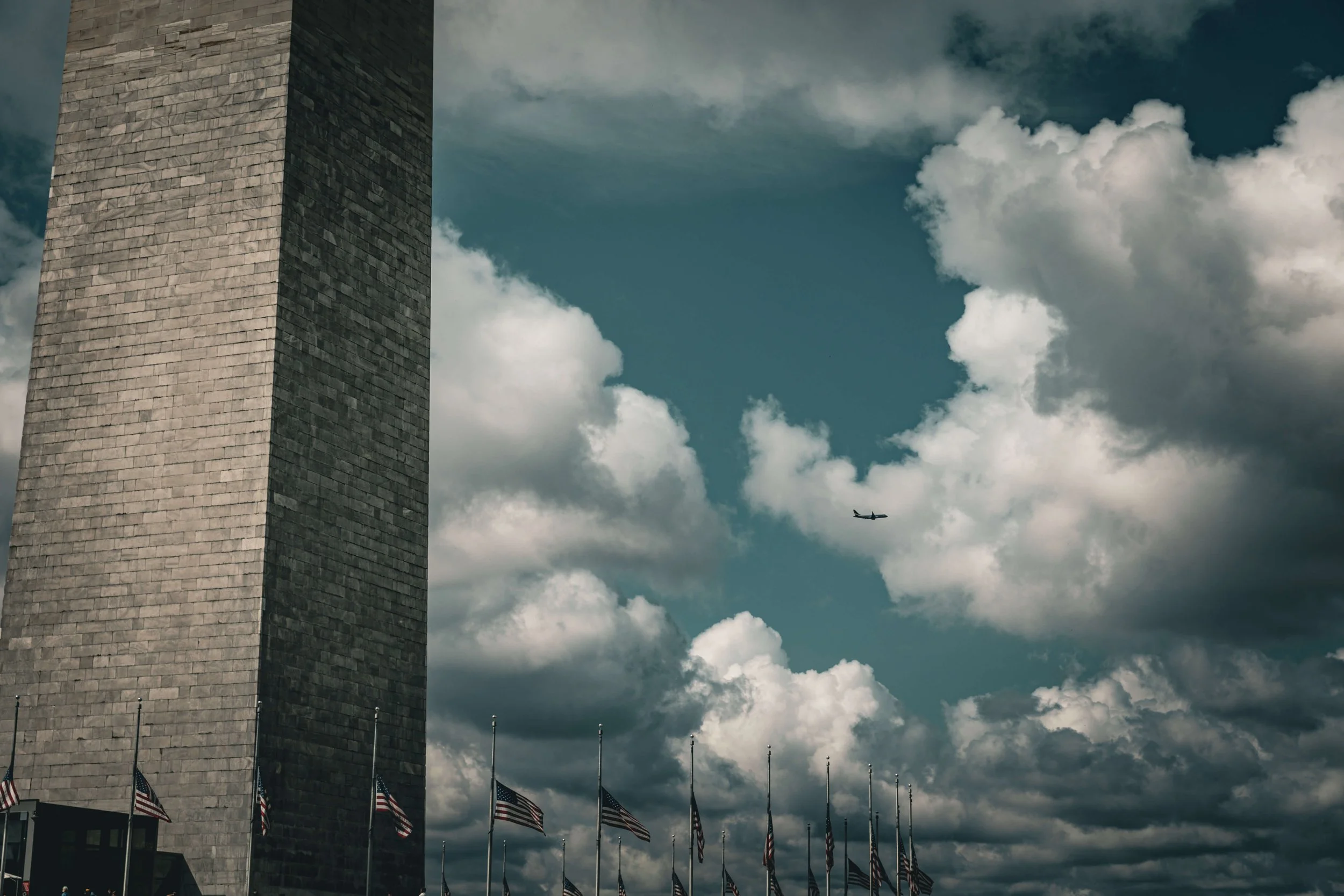 The Washington Monument towering over American flags with an airplane flying through a cloudy sky.