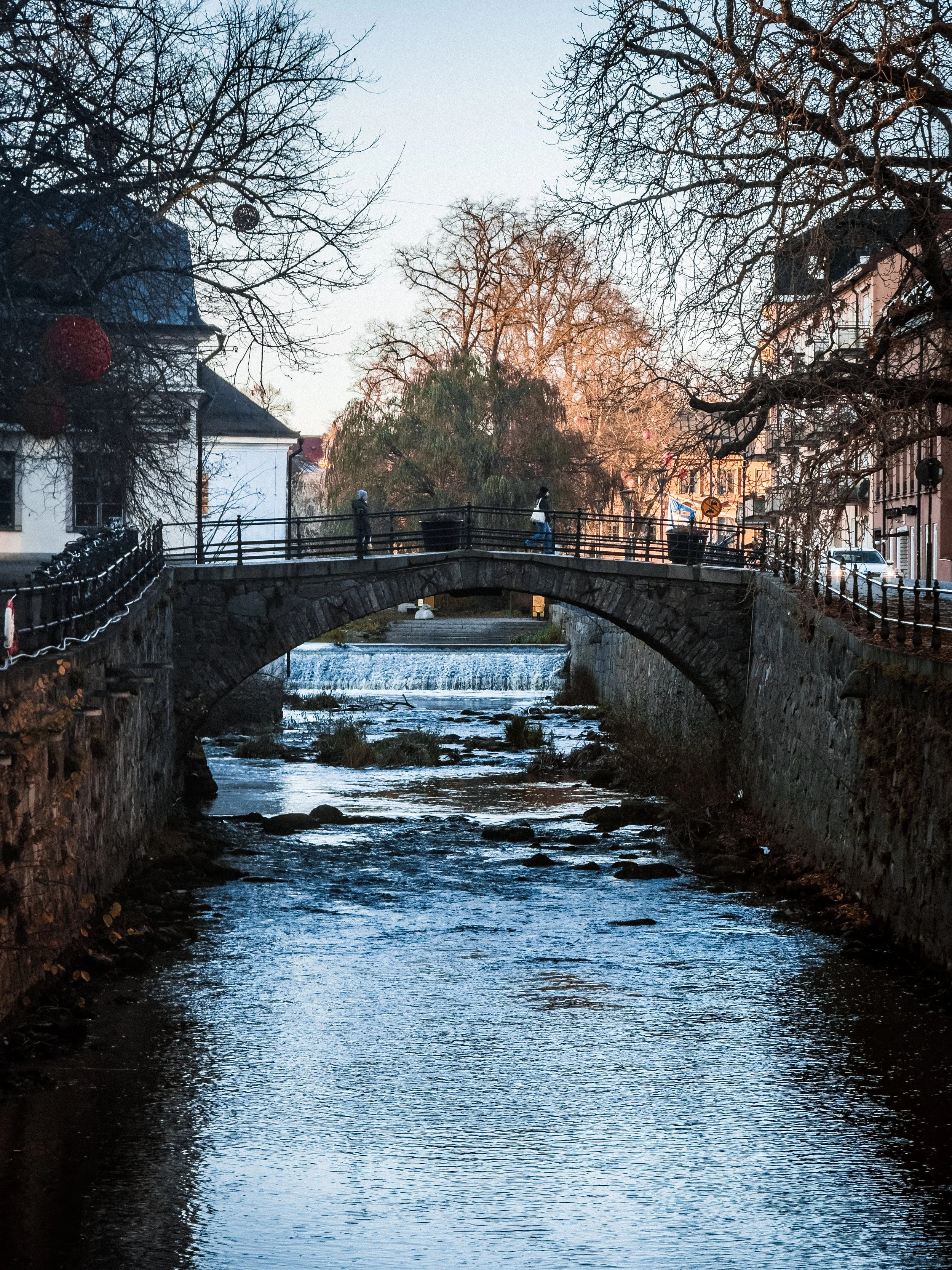 A small stone bridge over a river with flowing water, bordered by stone walls and leafless trees, in a city with buildings on either side, during sunset or late afternoon.