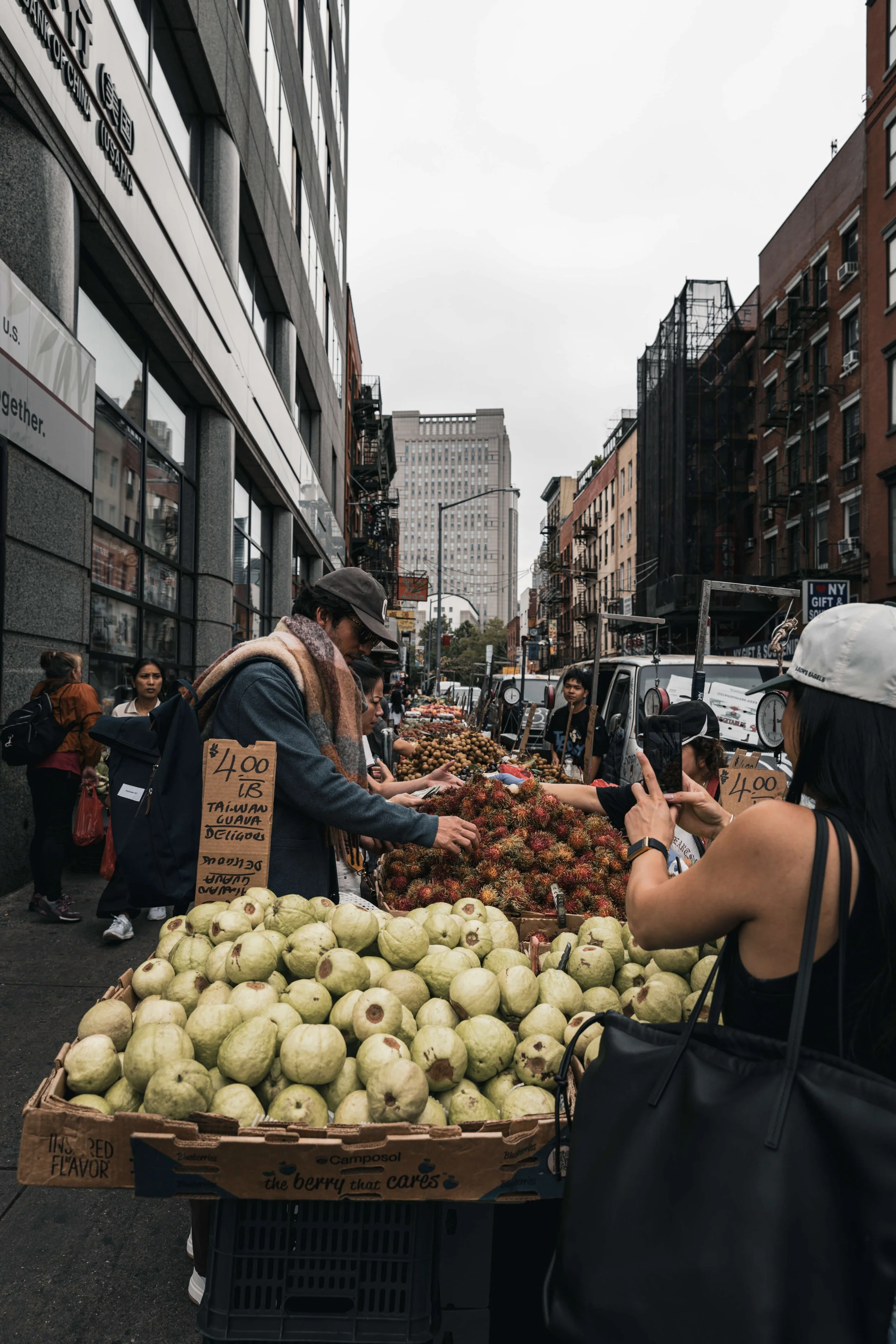Street market scene with vendors selling fresh fruits including guavas and rambutans, and a woman taking a photo of the market with her phone.