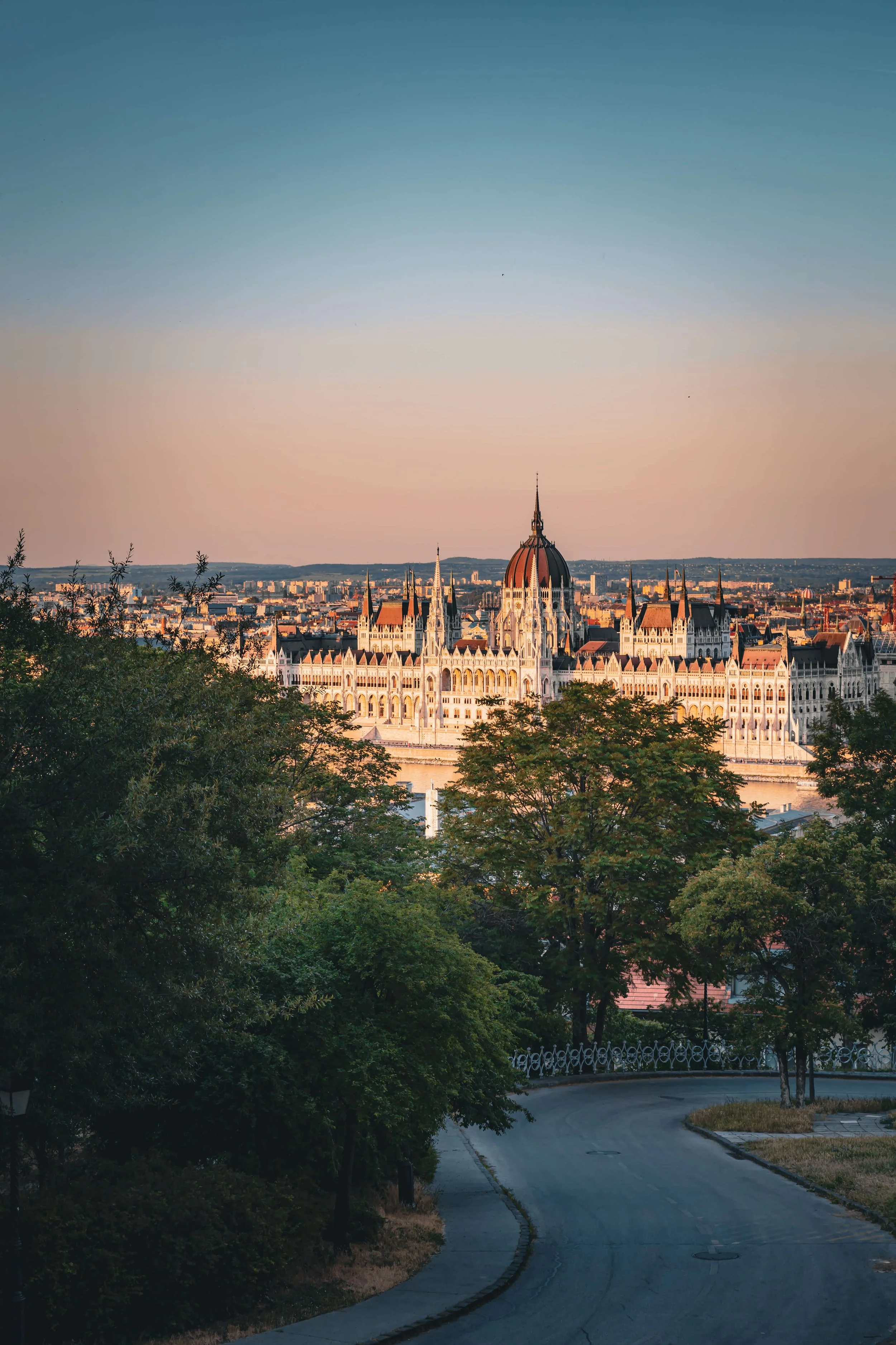 A view of the Hungarian Parliament Building in Budapest, Hungary, seen from a park with trees in the foreground, during sunset.