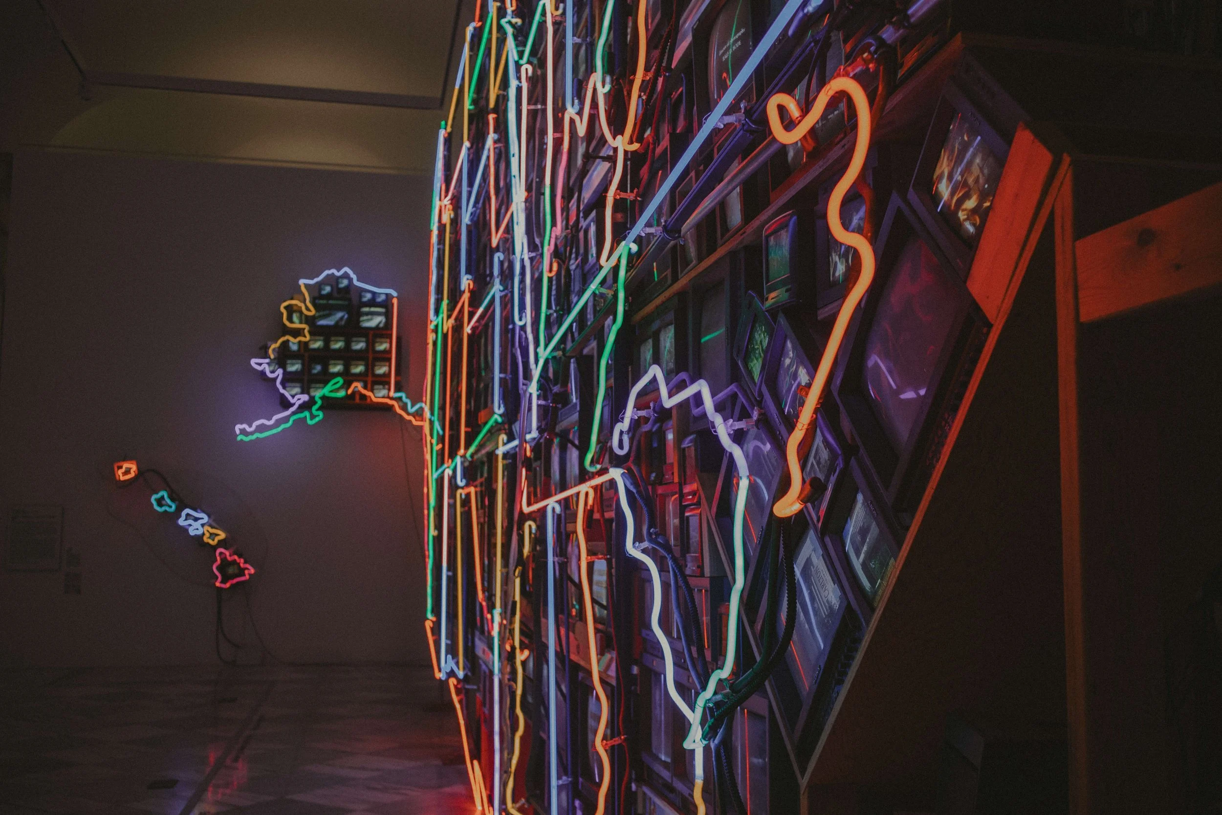 Colorful neon light art installation depicting a map outline with various neon colors, mounted on a display with small television screens