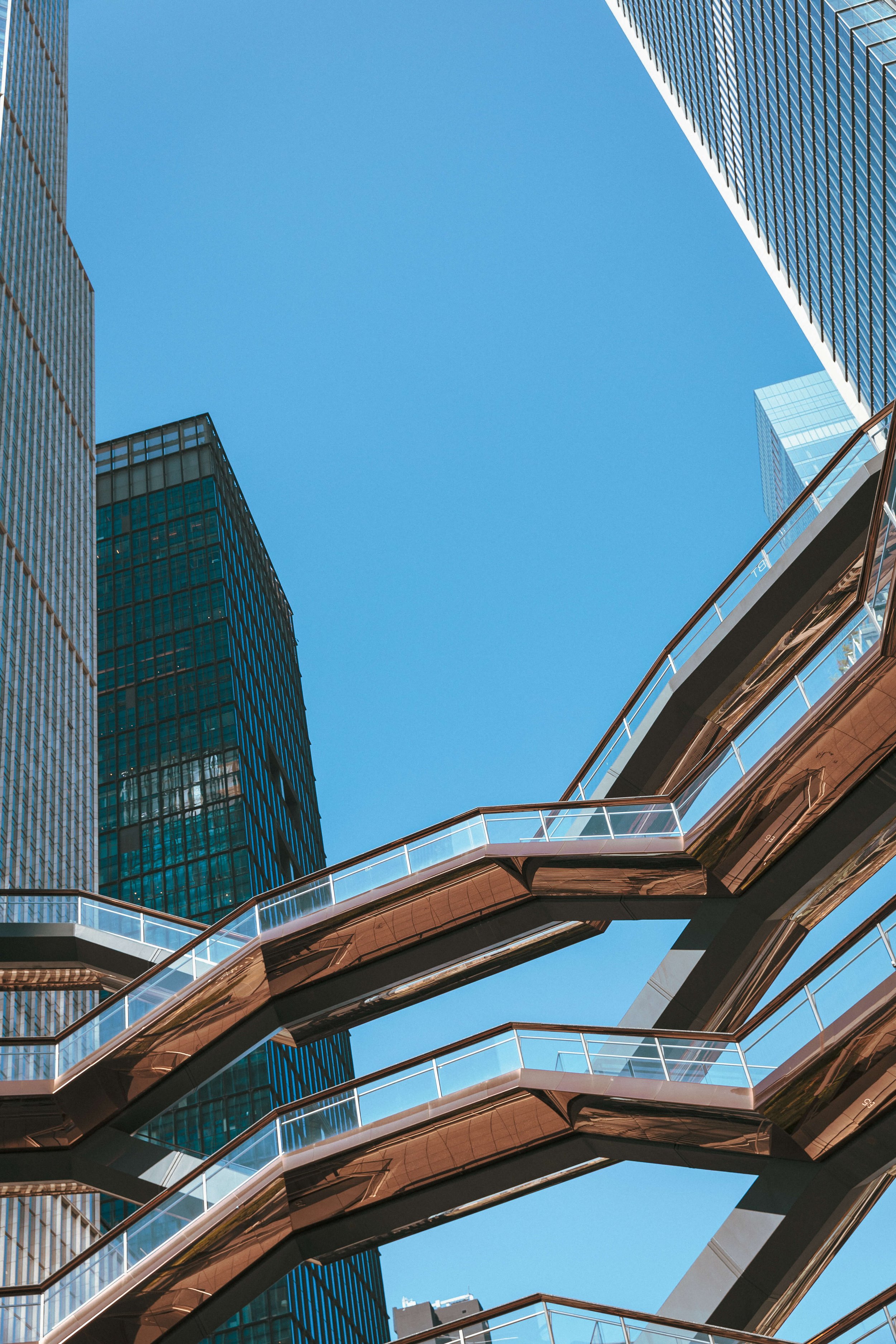 View of modern high-rise buildings and a spiral pedestrian bridge against a bright blue sky.