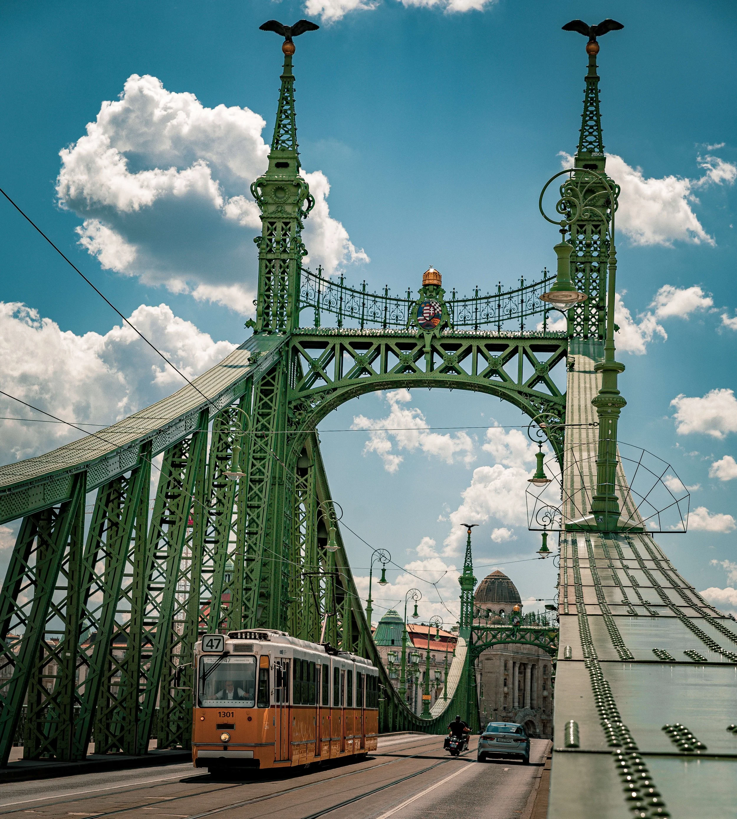 A large green bridge with intricate ironwork, featuring two towers topped with bird sculptures. A tram is traveling along the bridge, and there are cars and motorcycles below. The bridge is set against a backdrop of a blue sky with scattered white cl