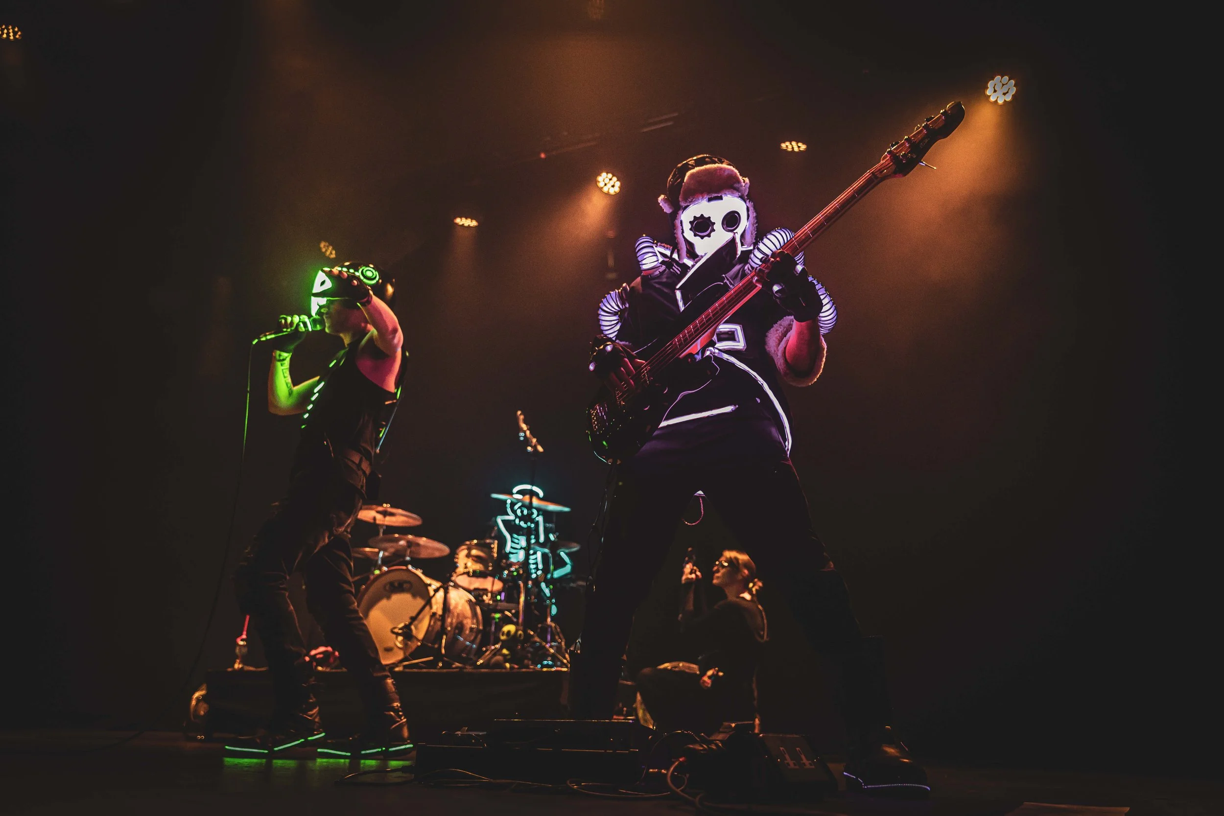 A band performs on stage wearing futuristic costumes with glowing features, and a skeleton design on the drum set.