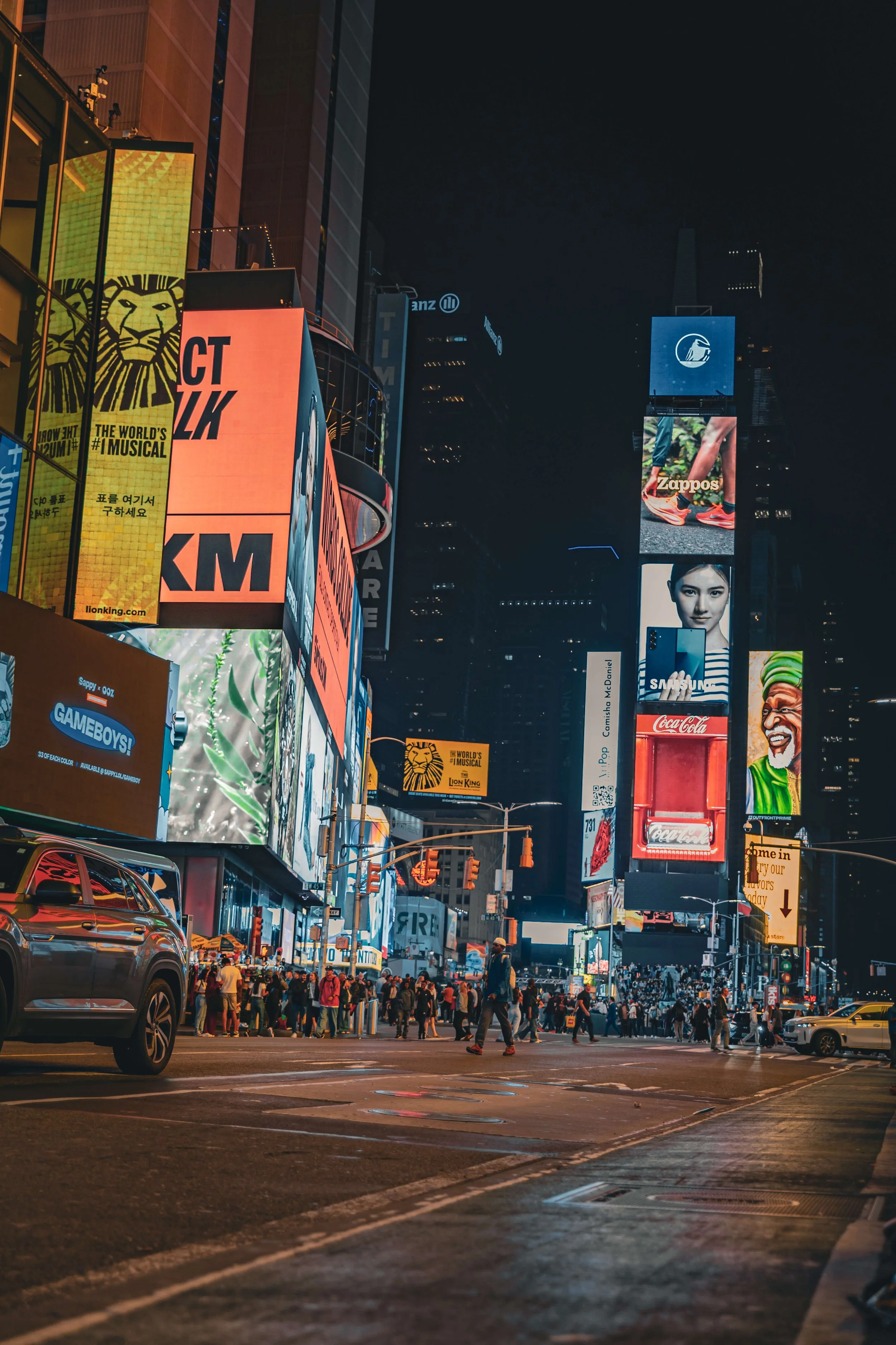 Nighttime view of Times Square, New York City, with illuminated billboards, advertisements, and a crowd of pedestrians crossing the street.
