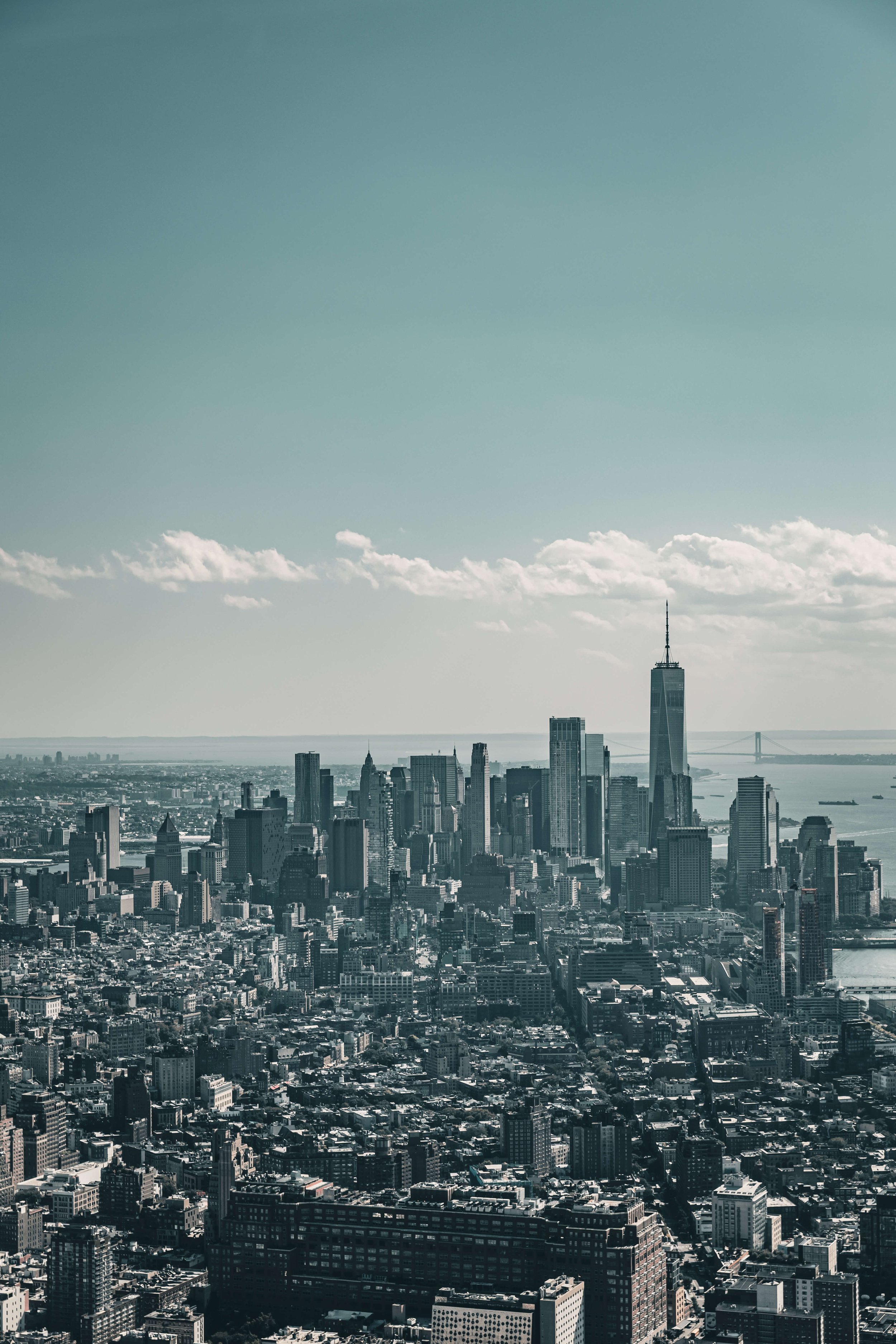 Aerial view of New York City skyline with skyscrapers including One World Trade Center, and the Brooklyn Bridge in the distance under a clear sky.