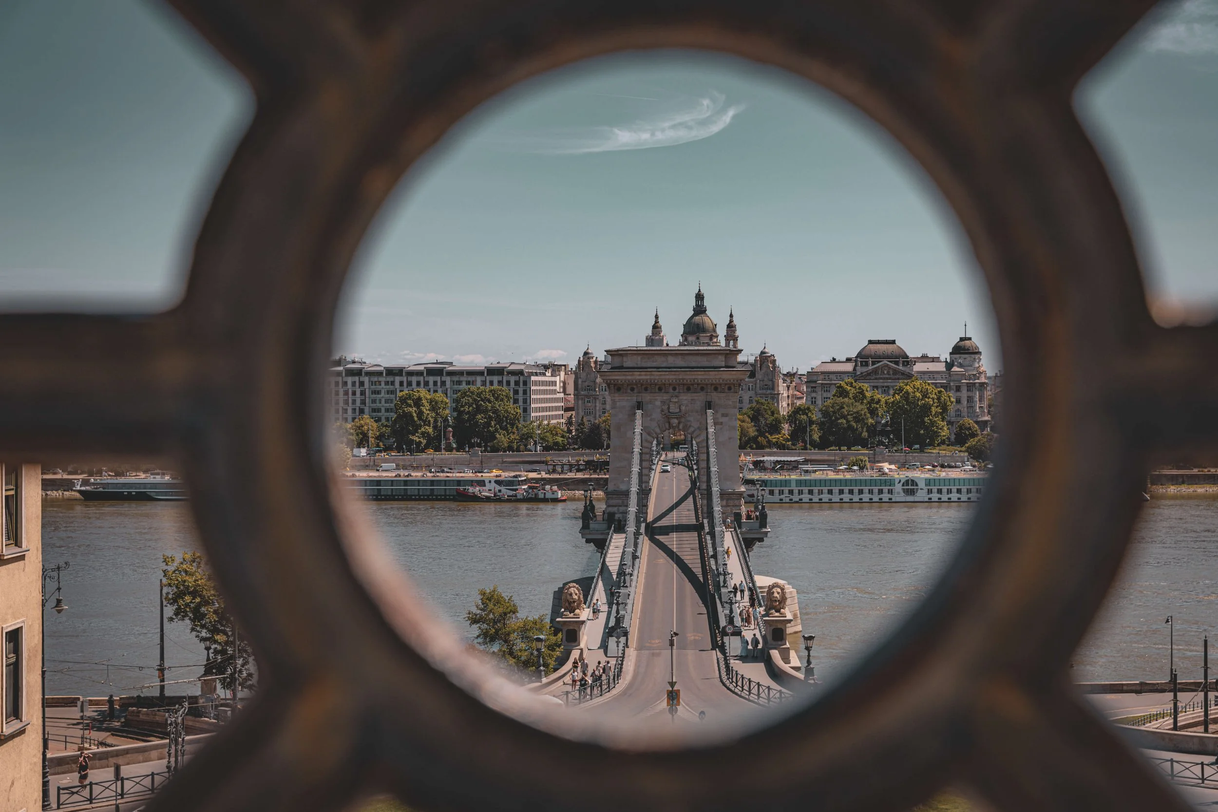 View of a cityscape with a bridge crossing a river, seen through a decorative iron railing, with buildings and a church with domes in the background, under a partly cloudy sky.