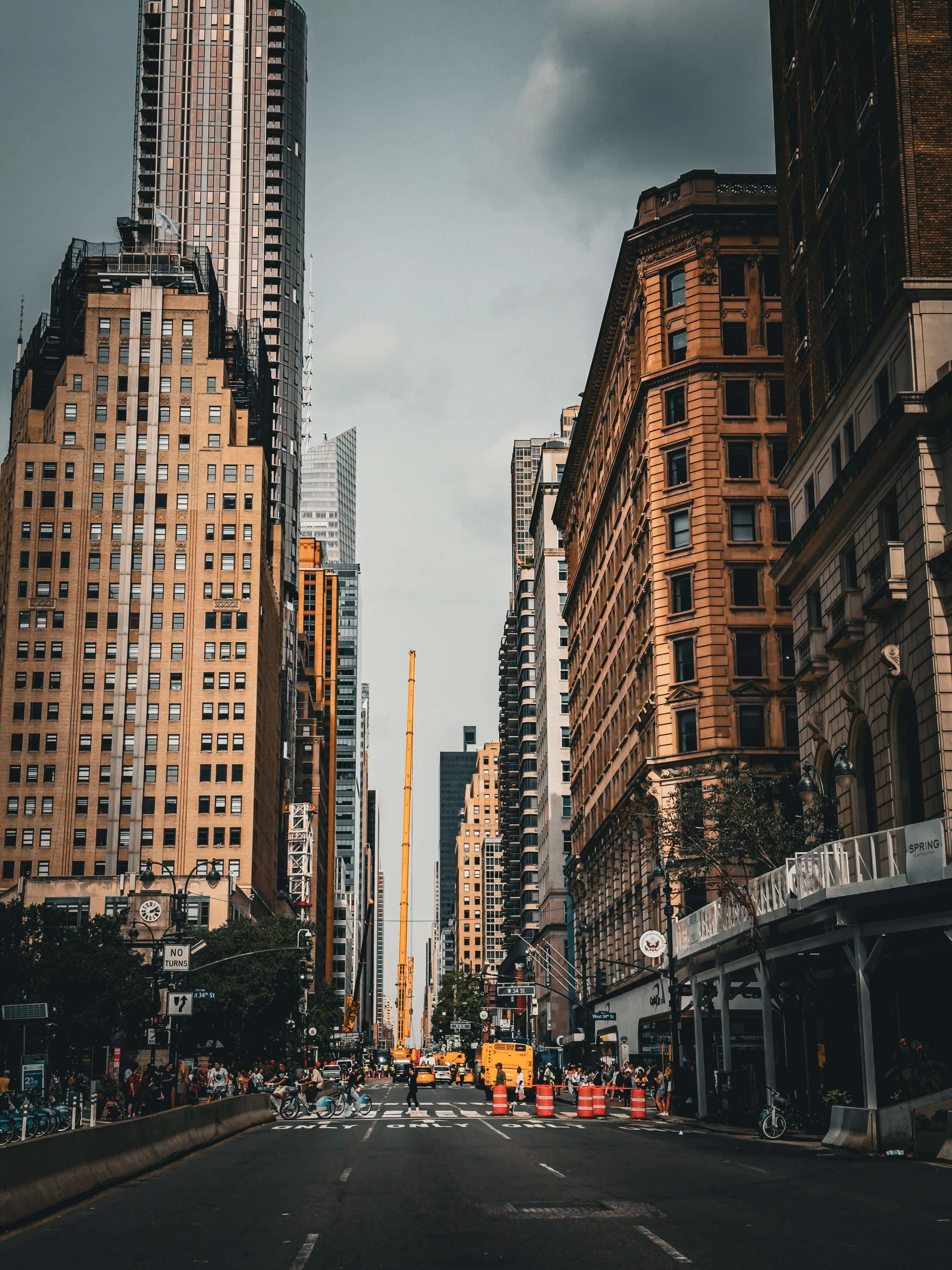 Urban street scene in a city, with tall skyscrapers on both sides, pedestrians crossing, construction cones, and a yellow taxi in the distance.