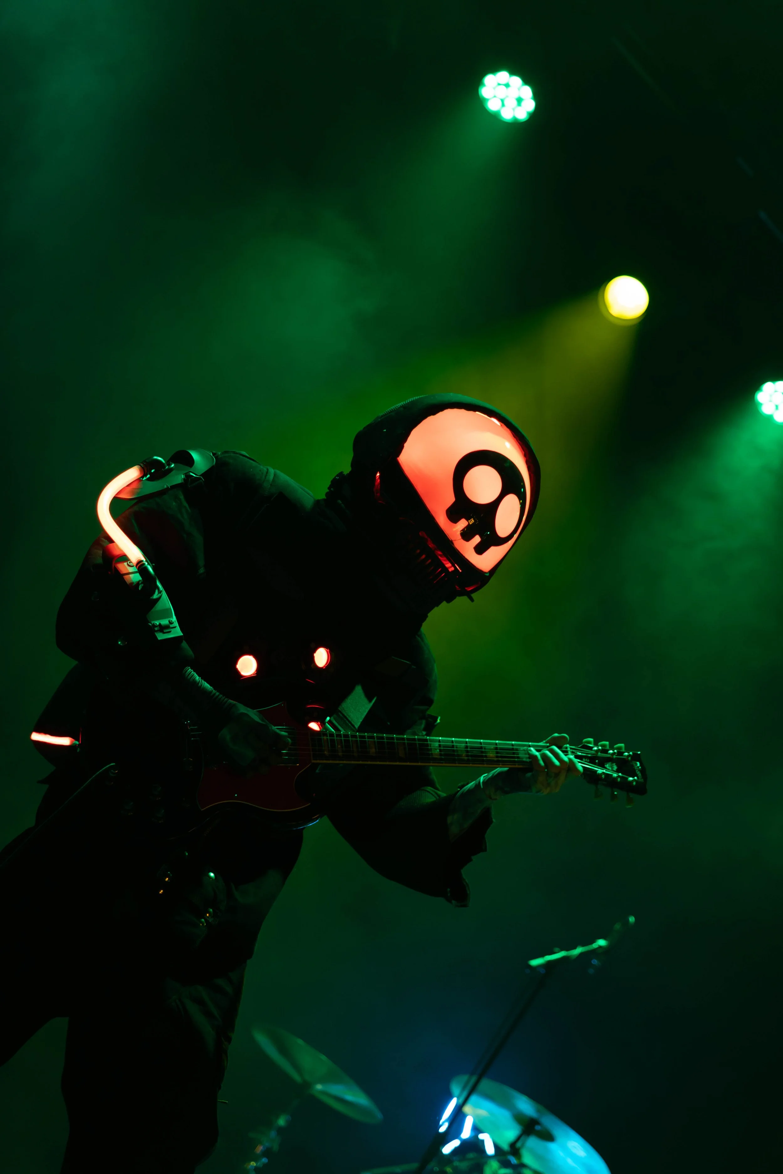 A musician dressed in black, wearing a glowing skull mask, playing an electric guitar on stage illuminated by green lights.