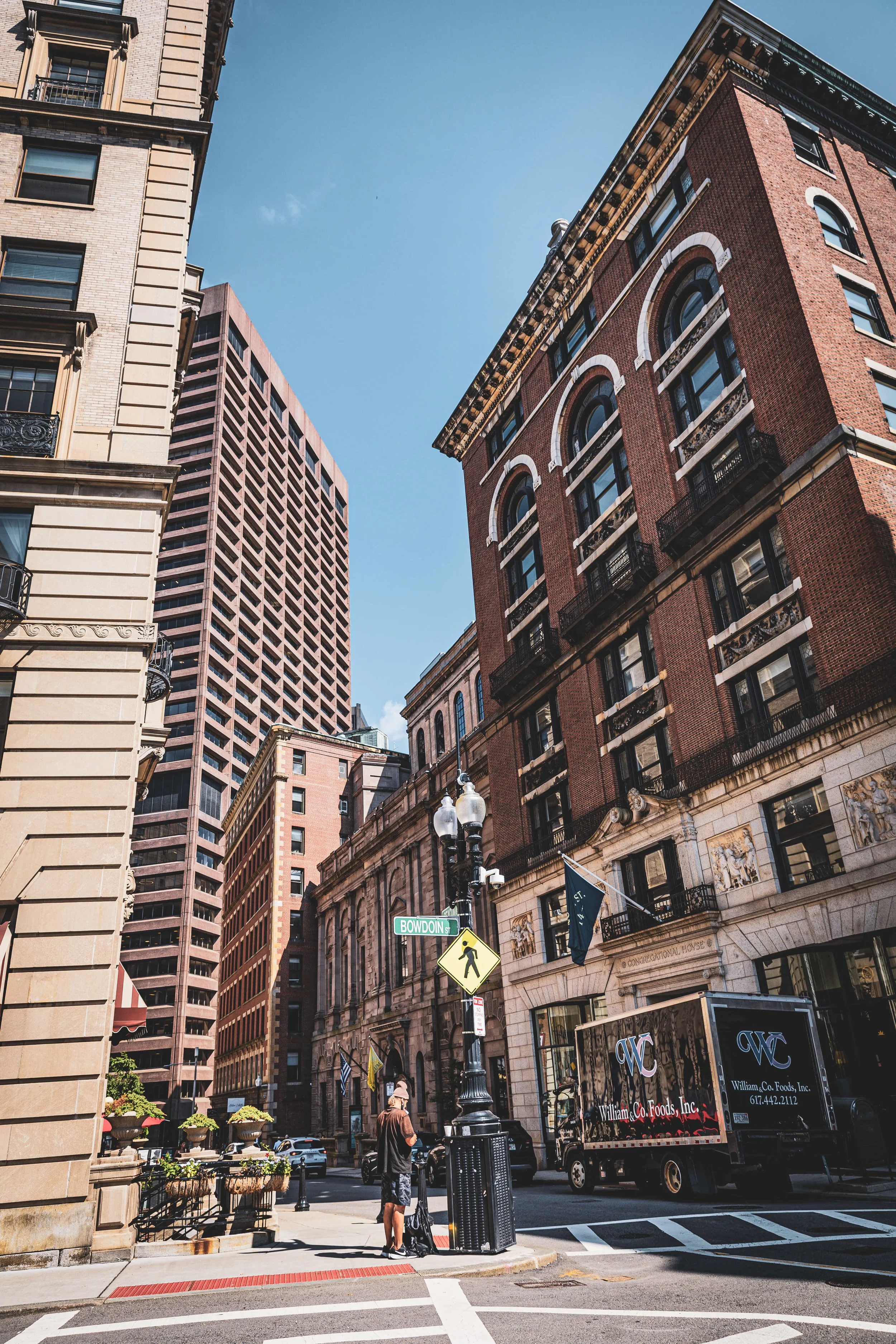 City street scene with tall buildings, a man walking with luggage, street signs for Bowdoin Street, and a William Co. Foods delivery truck.