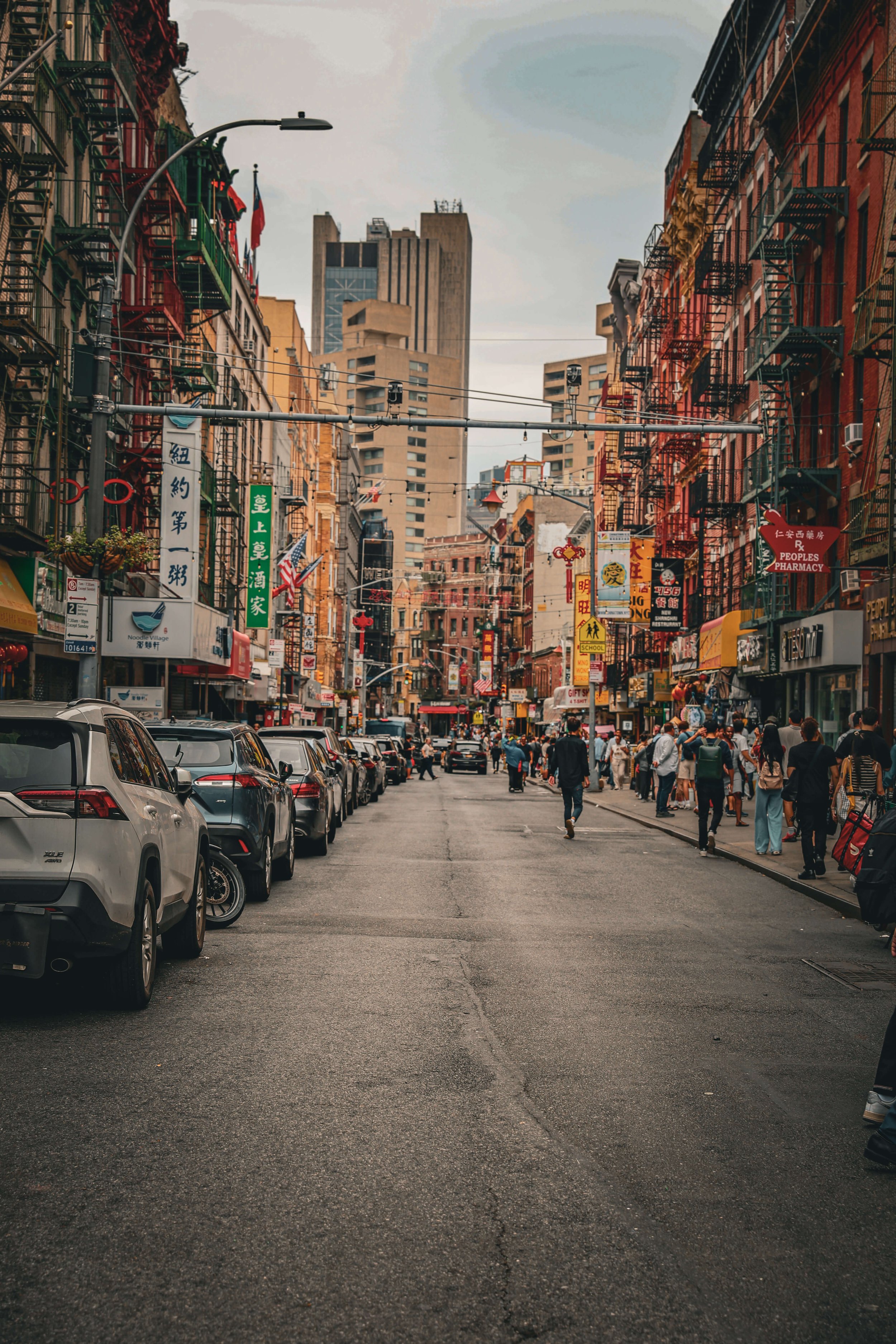 A busy city street with cars parked along the sides, people walking on the sidewalks, and colorful signs in Chinese characters. Tall buildings are visible in the background under a cloudy sky.