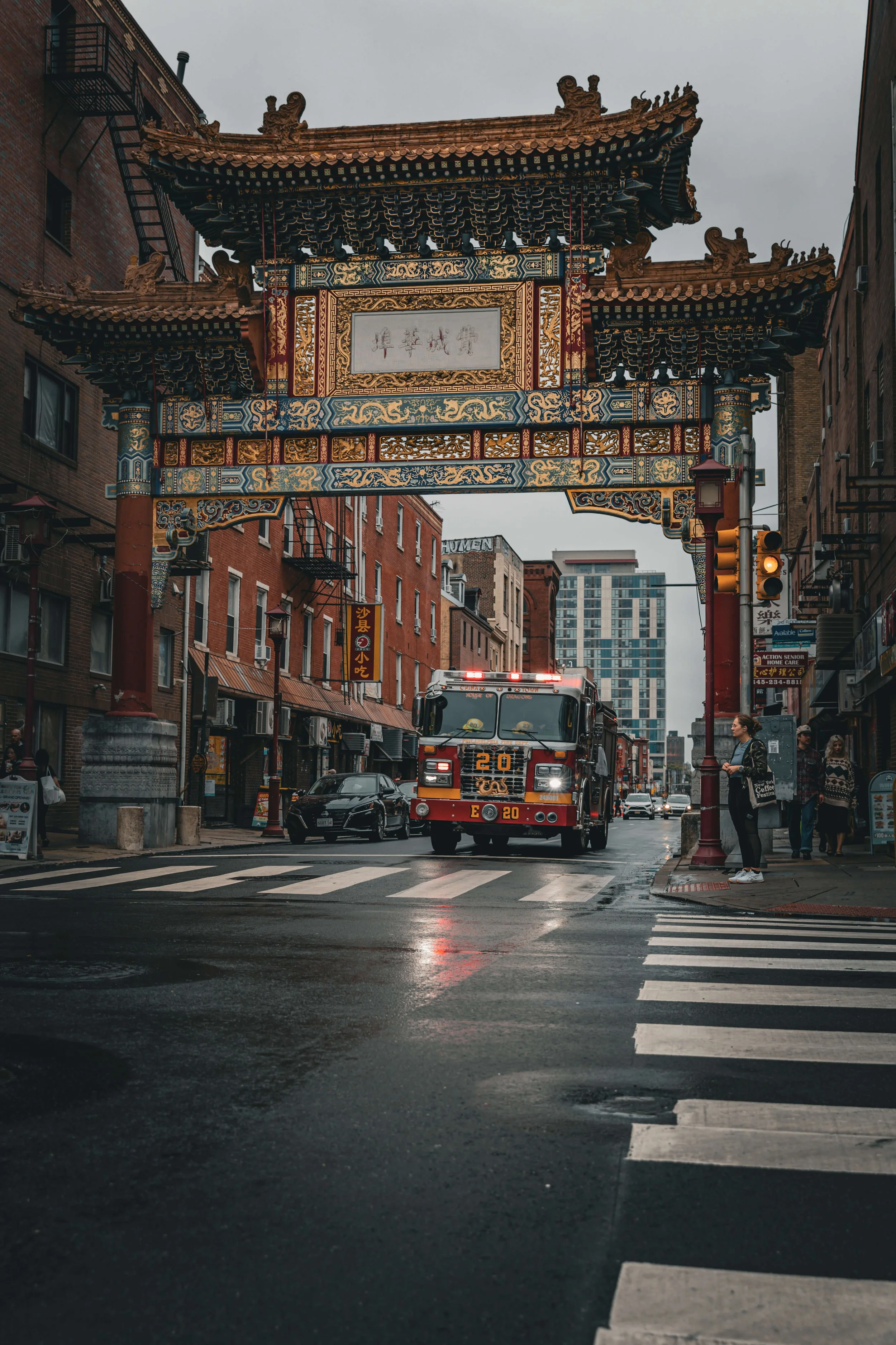 Street scene in Chinatown with a decorative Chinese archway overhead, a fire truck crossing at an intersection, and pedestrians waiting on the sidewalk.