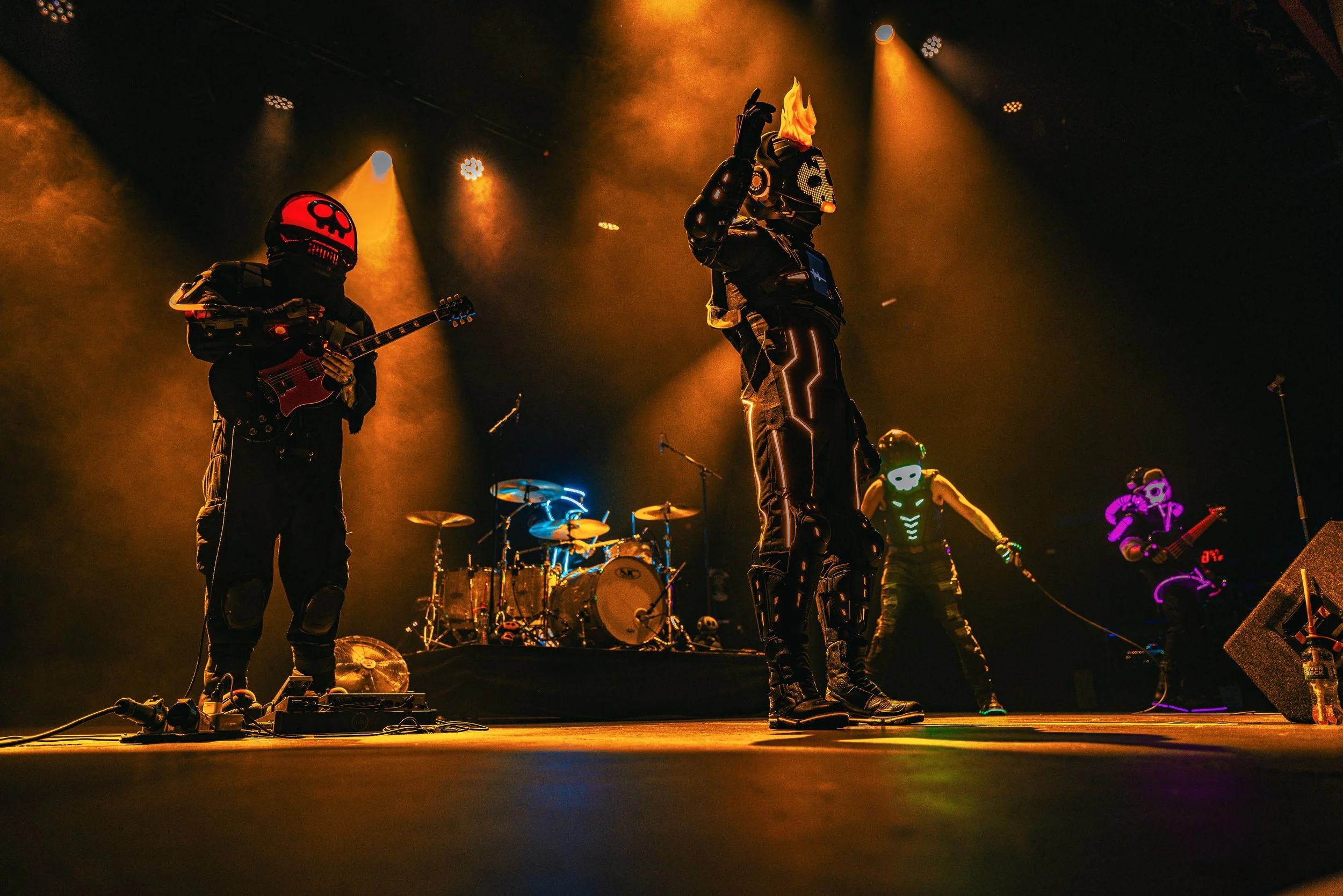 A band performs on stage with neon lights and smoke effects. The lead singer is wearing a suit with illuminated lines and a mask with a flame design, holding a microphone. The guitarist and other band members are dressed in dark, futuristic costumes 