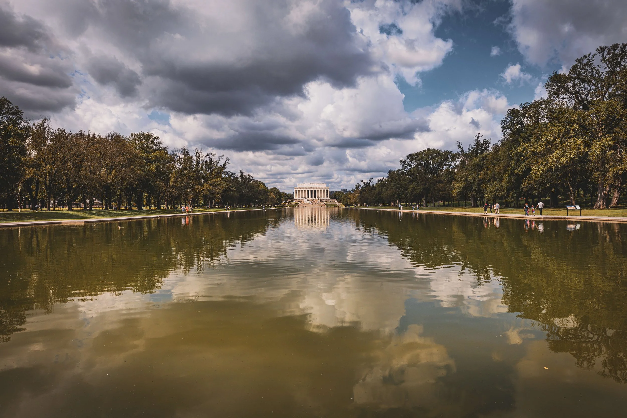 Washington Monument reflected in the Reflecting Pool with trees on either side and cloudy sky above.