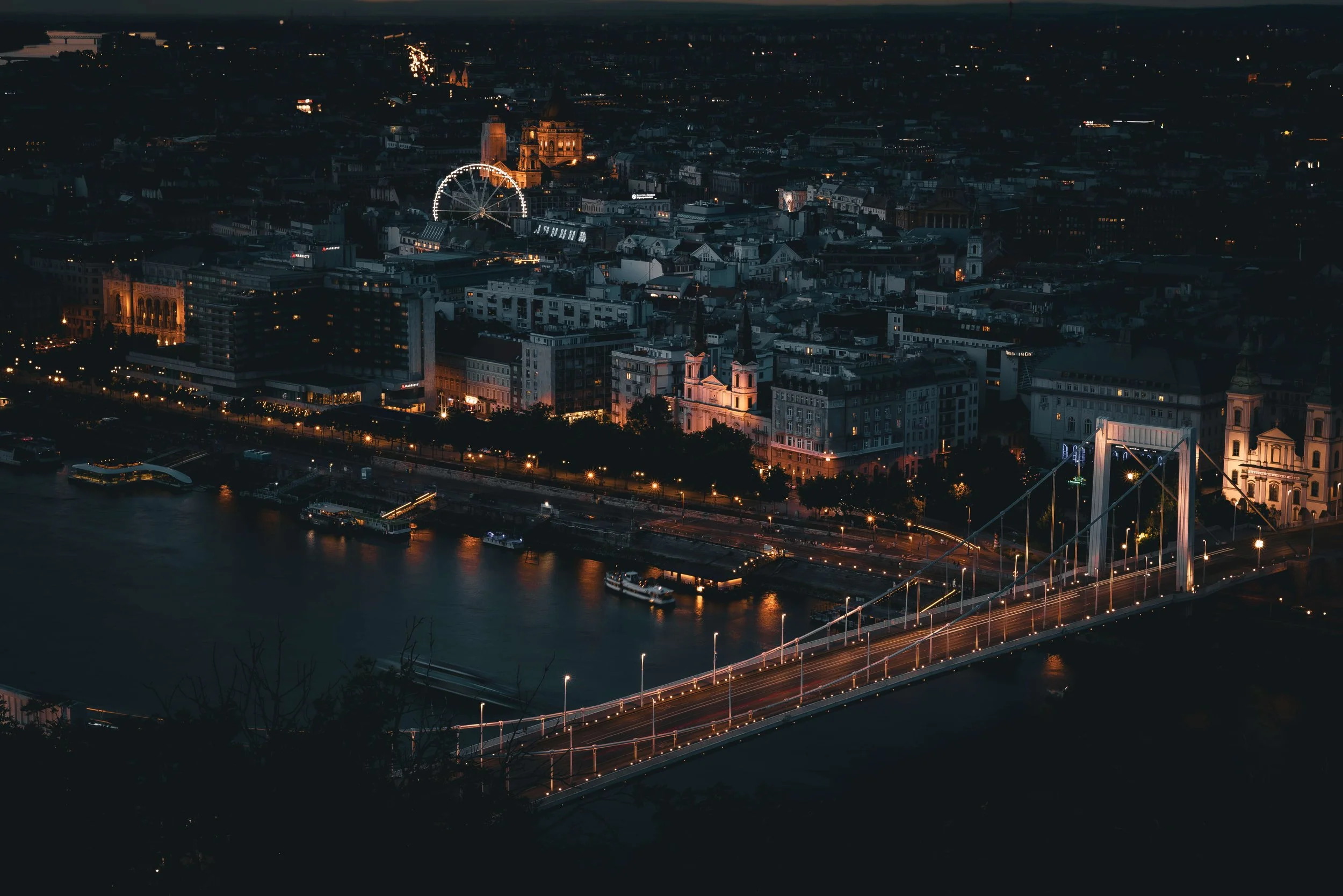 Nighttime aerial view of a cityscape with a river, illuminated buildings including churches, a Ferris wheel, and a bridge with light trails from passing vehicles.