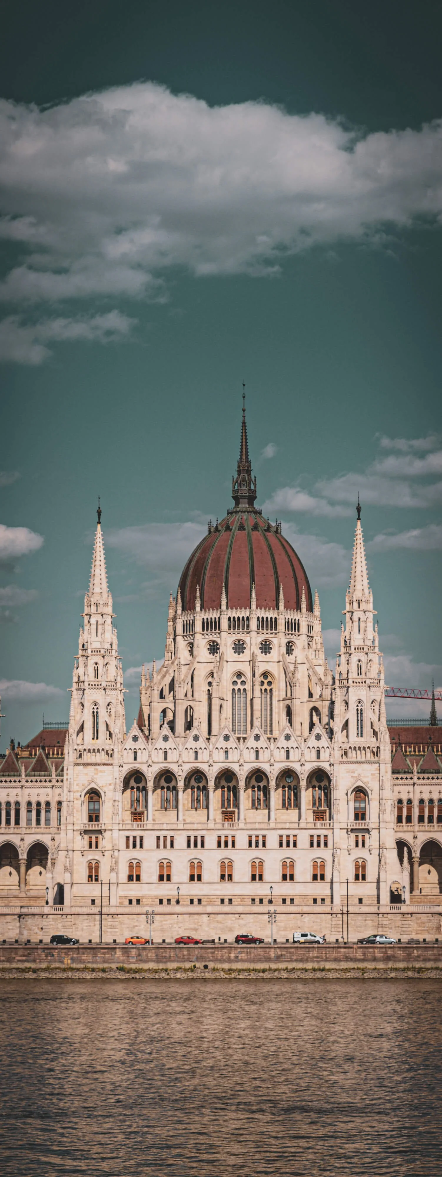 View of the Hungarian Parliament Building in Budapest, with its ornate neo-Gothic architecture, large central dome, and two towers, along the Danube River under a partly cloudy sky.