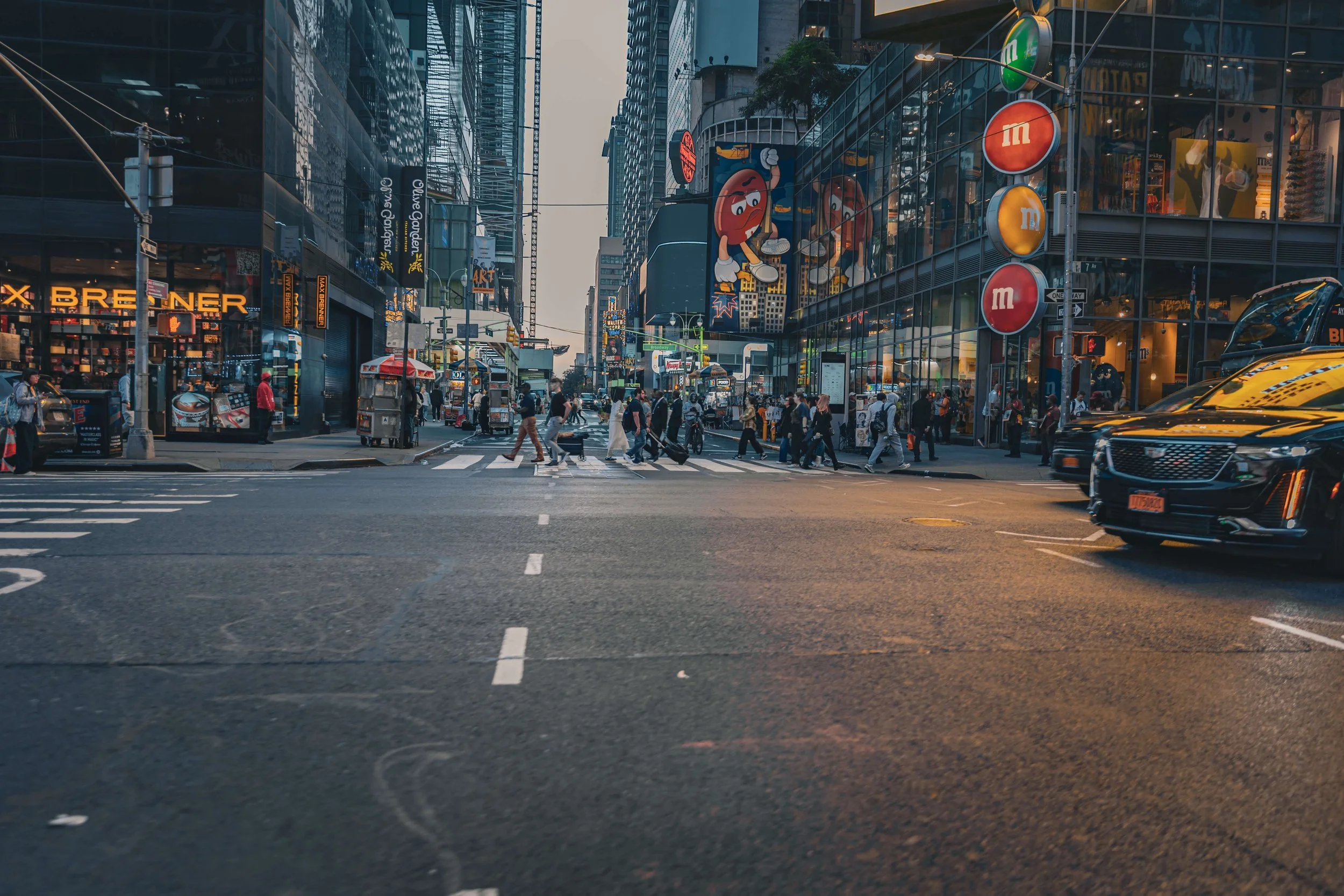 Busy city street with pedestrians crossing at a crosswalk. Tall buildings with illuminated signs and advertising on both sides of the street. A large animated billboard featuring a cartoon character and several M&M's logos are visible on the building