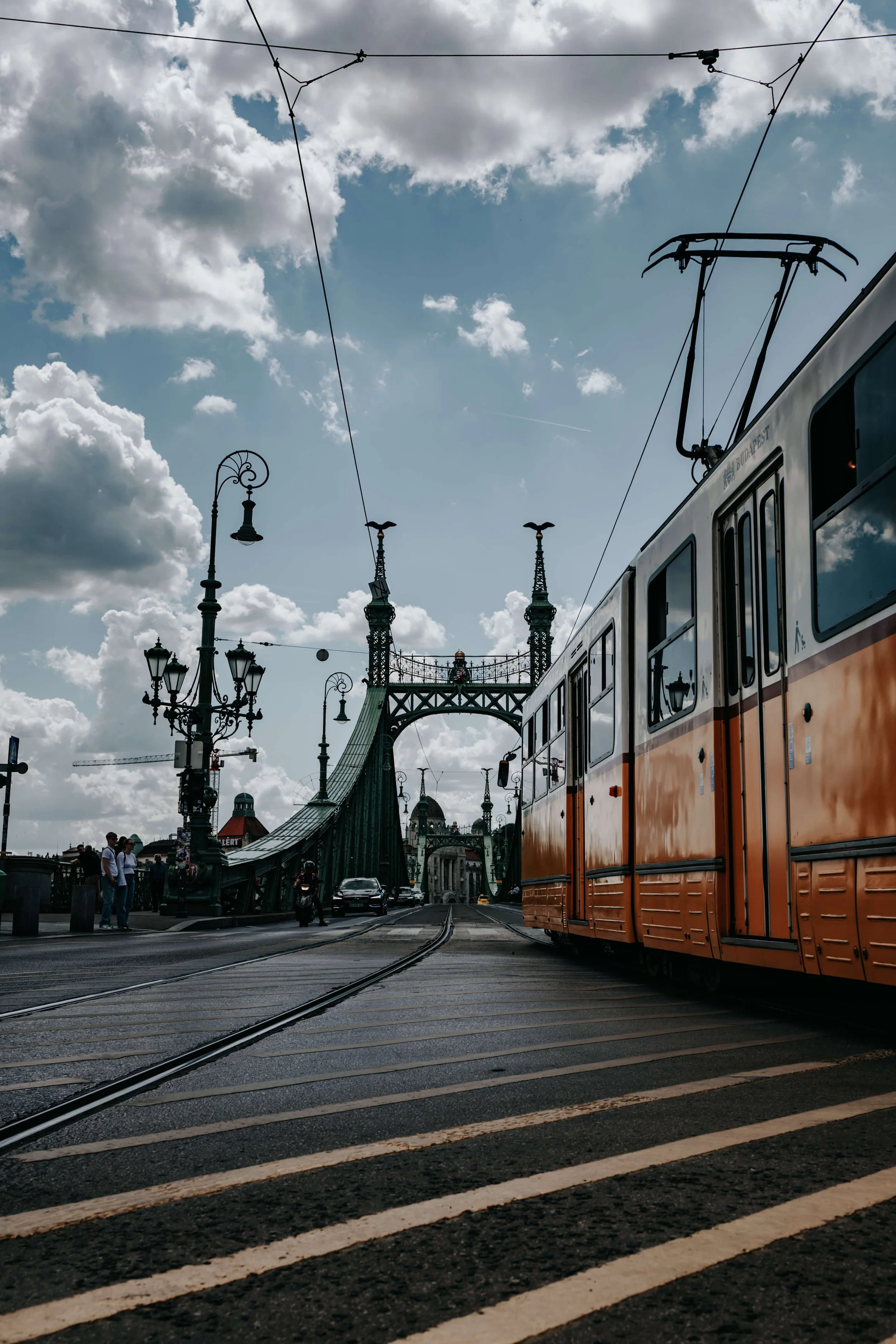 Orange tram on tracks crossing a historic bridge with pedestrians, street lamps, and city skyline under cloudy sky.