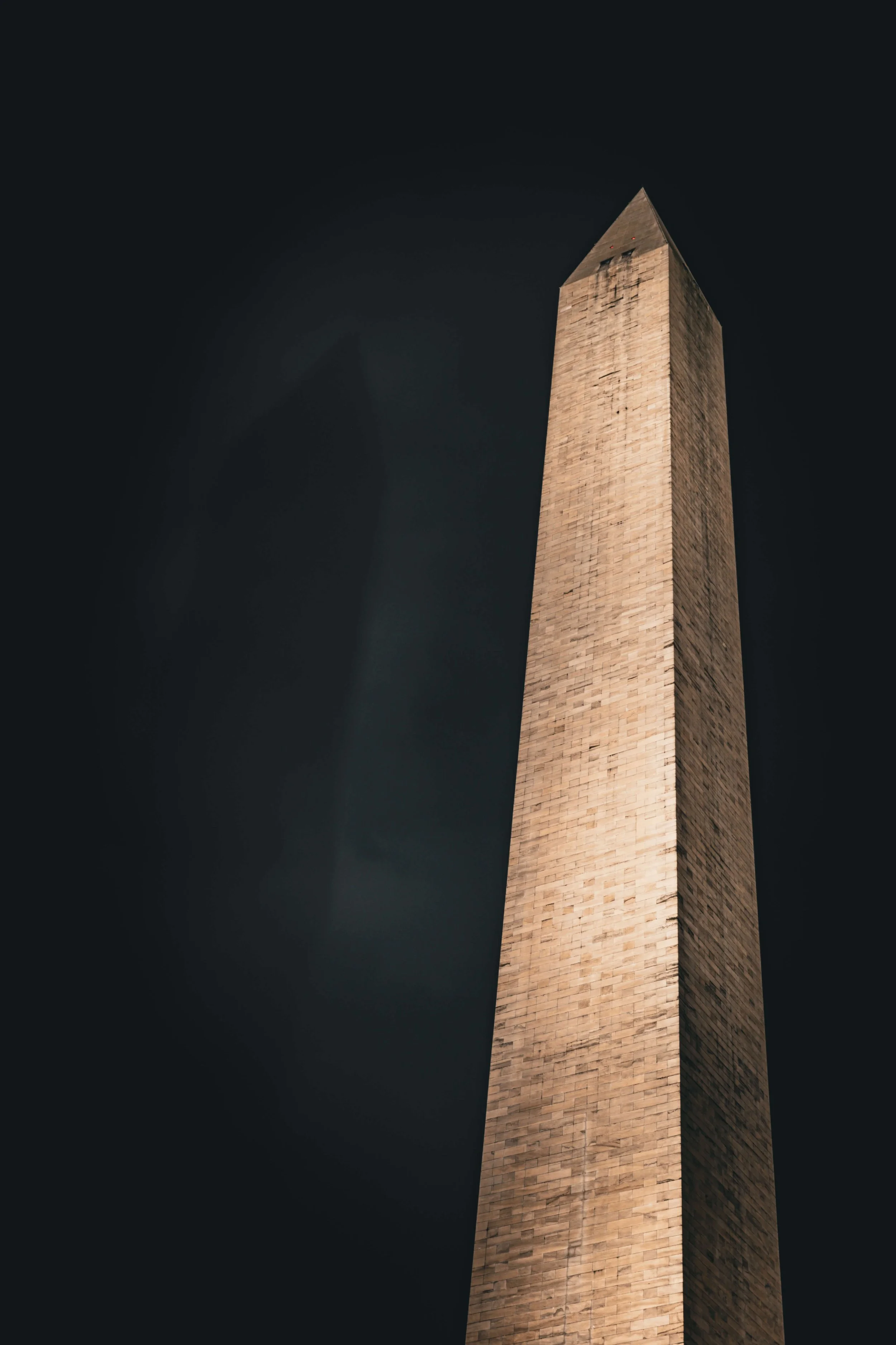 Nighttime view of a tall, rectangular stone obelisk with a pointed top, set against a dark sky.