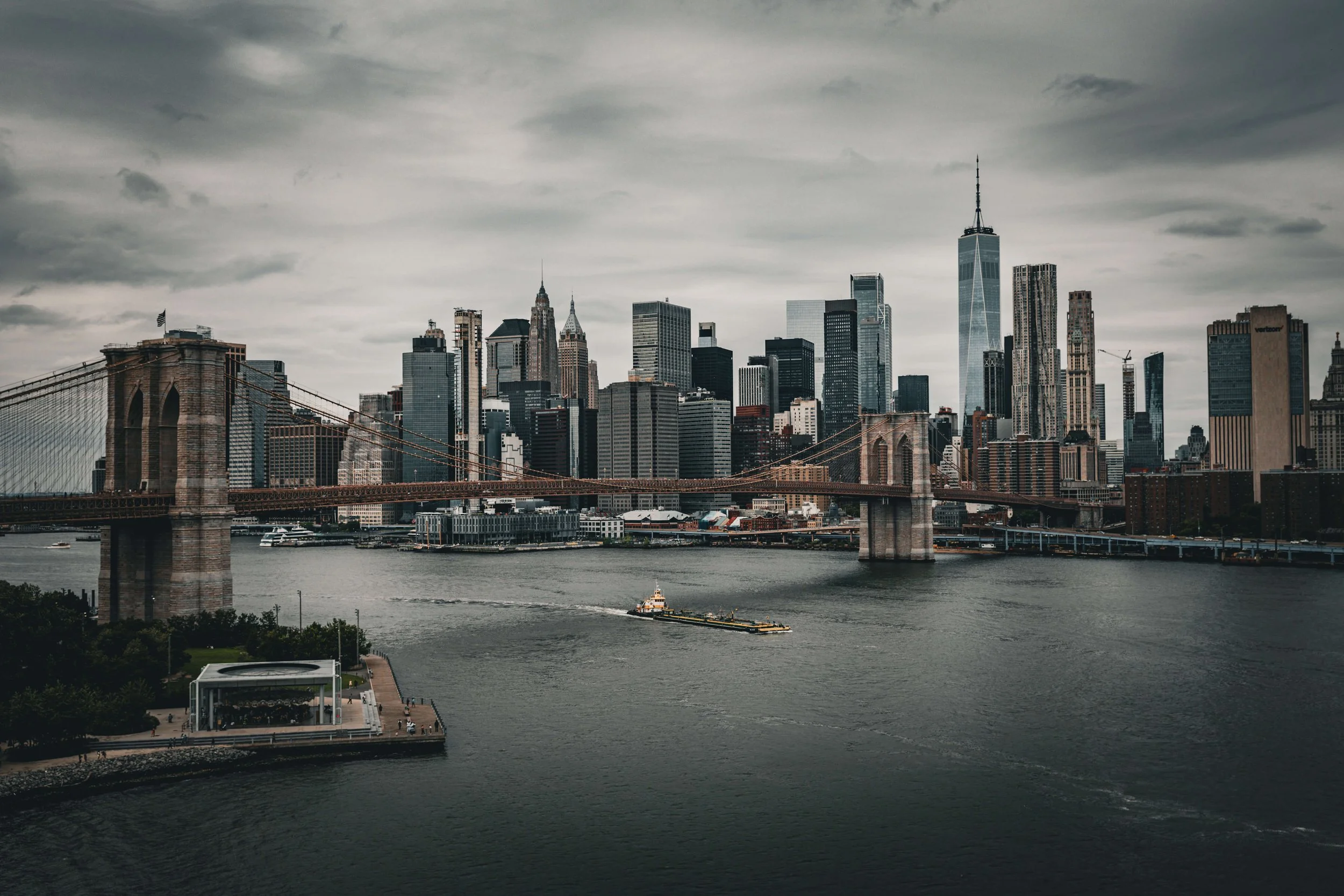 New York City skyline with Brooklyn Bridge and water in foreground, cloudy sky.