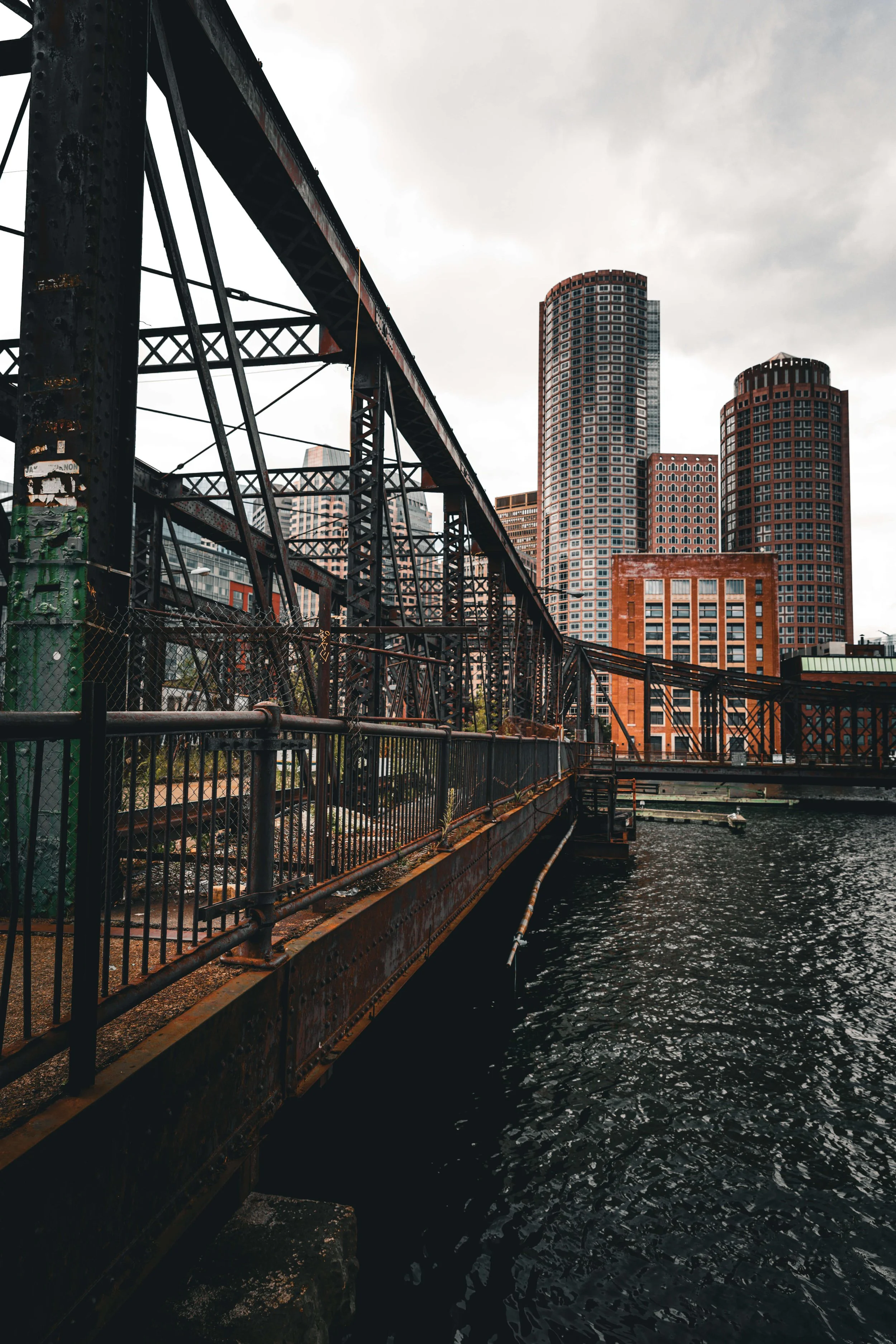Urban waterfront scene featuring an old rusted steel bridge with a city skyline of high-rise buildings in the background on a cloudy day.