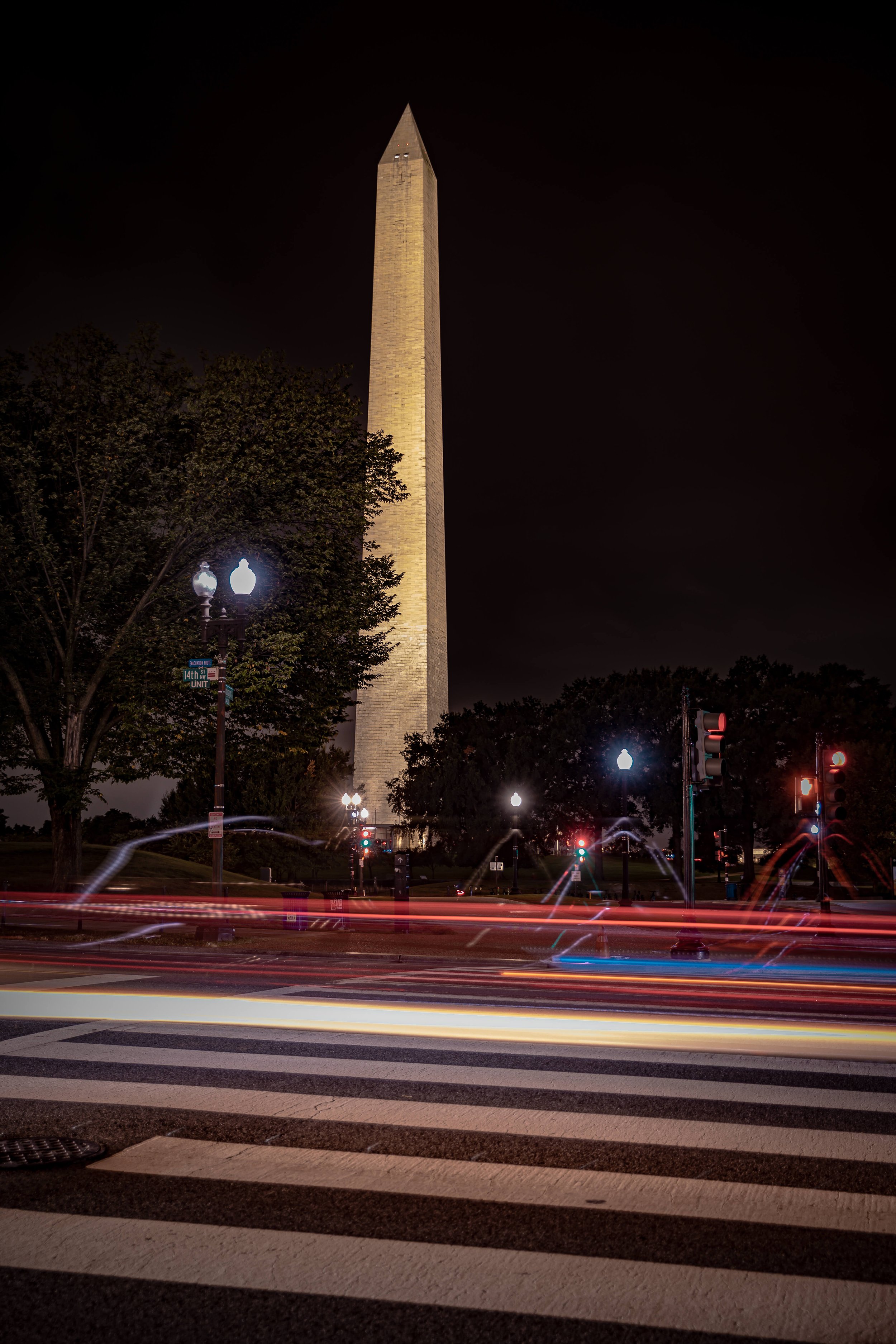 Nighttime view of the Washington Monument illuminated, with light trails from passing cars in the foreground and streetlights and trees surrounding the monument.