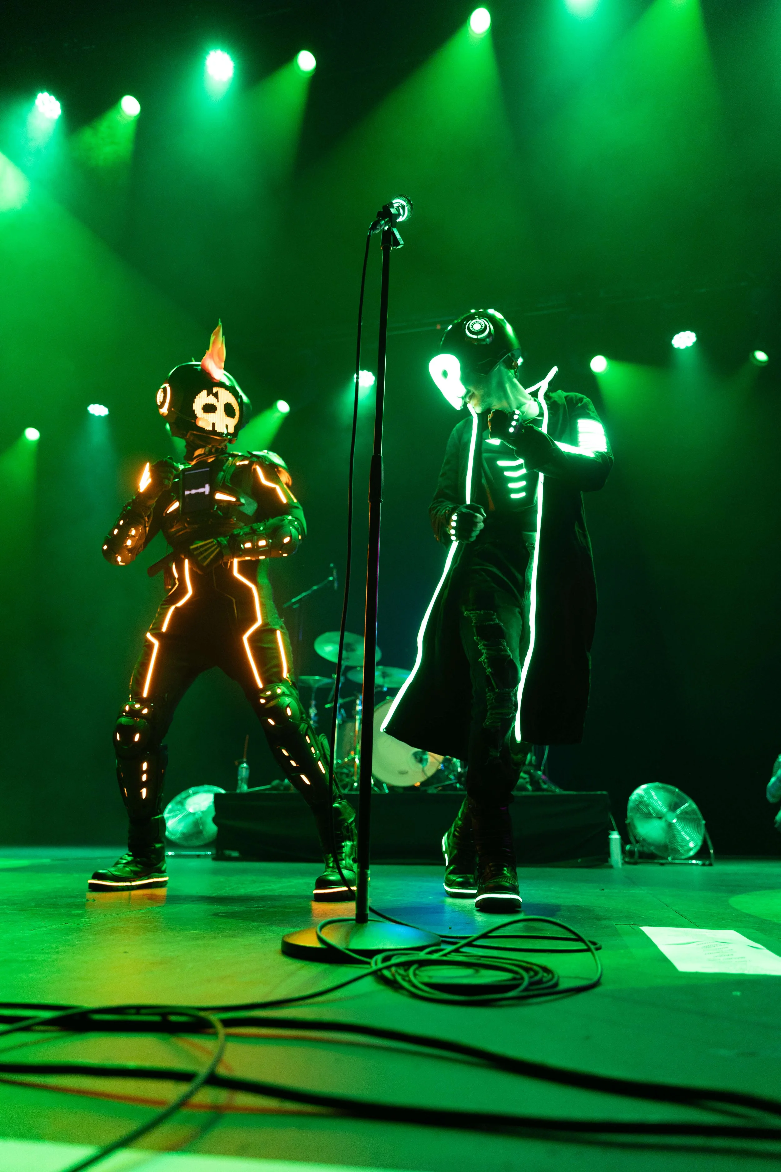 Two performers dressed in glowing, neon-outlined costumes on stage under green lighting, with microphones and cables in the foreground.
