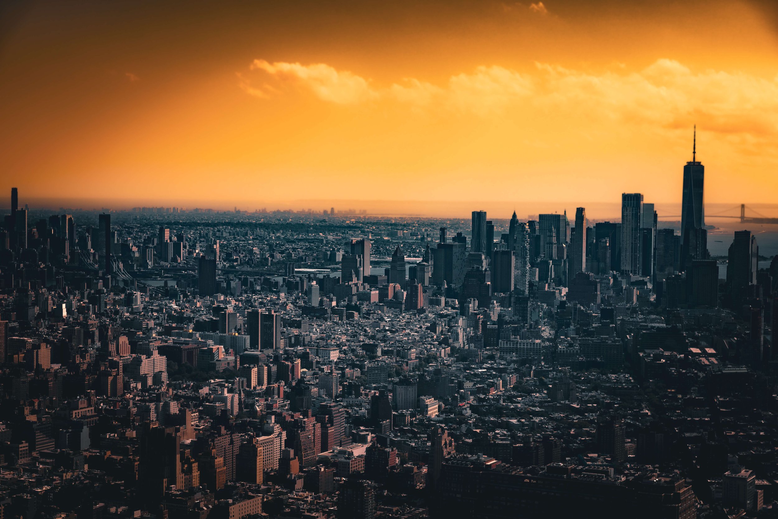 Skyline of New York City with tall skyscrapers and a bridge in the background during sunset with orange clouds.