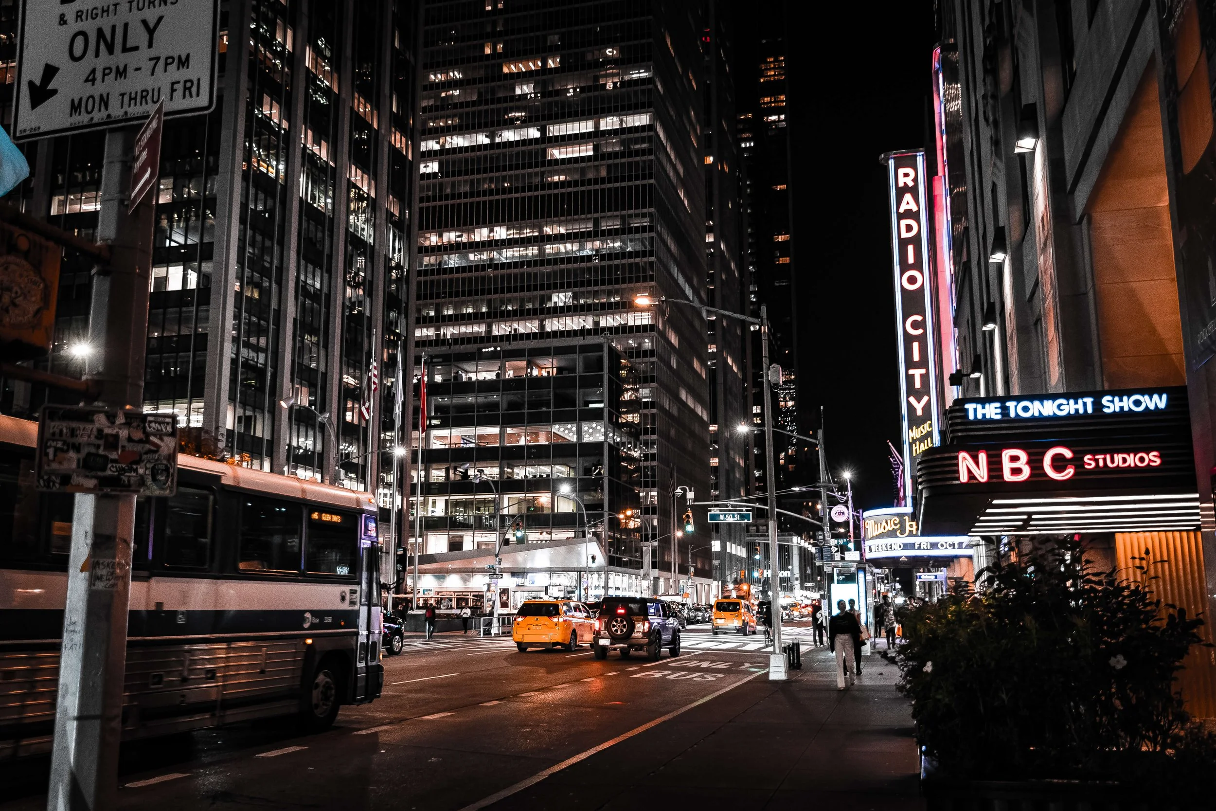 Nighttime city street scene with tall buildings, illuminated signs including Radio City, NBC Studios, and The Tonight Show, a bus, taxis, and pedestrians.