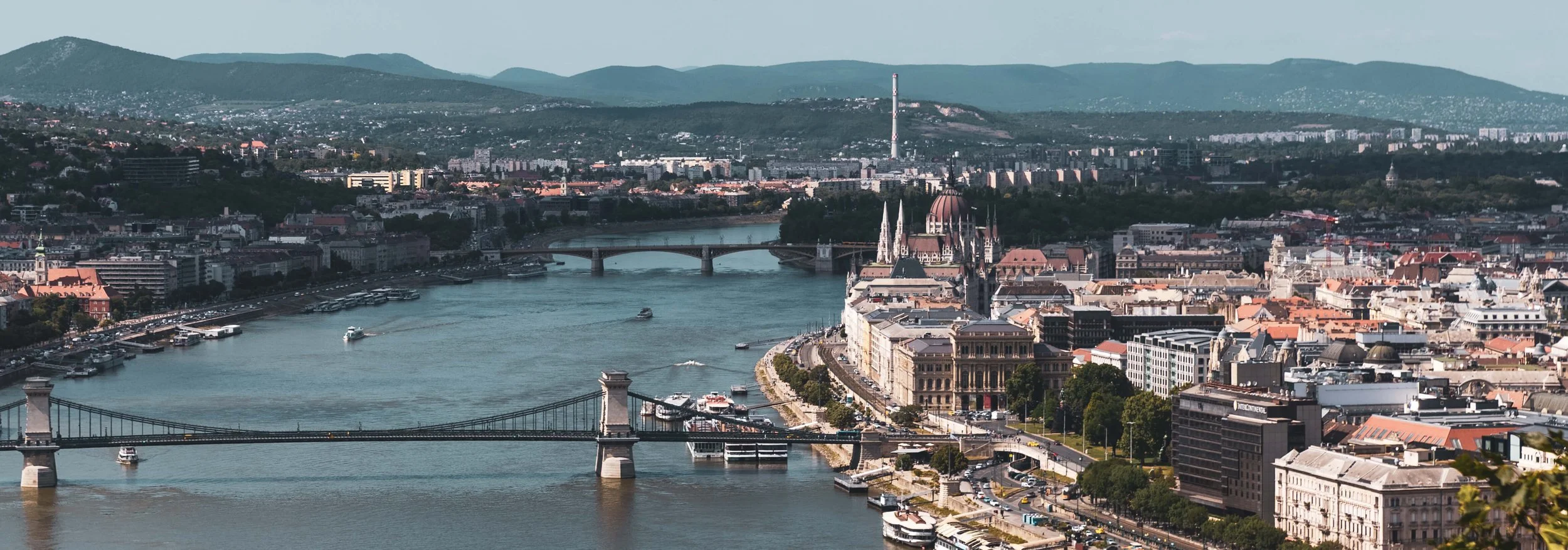 A panoramic view of Budapest, Hungary, featuring the Chain Bridge over the Danube River, the Parliament building with its distinctive dome, and Buda Castle, with mountains in the background.