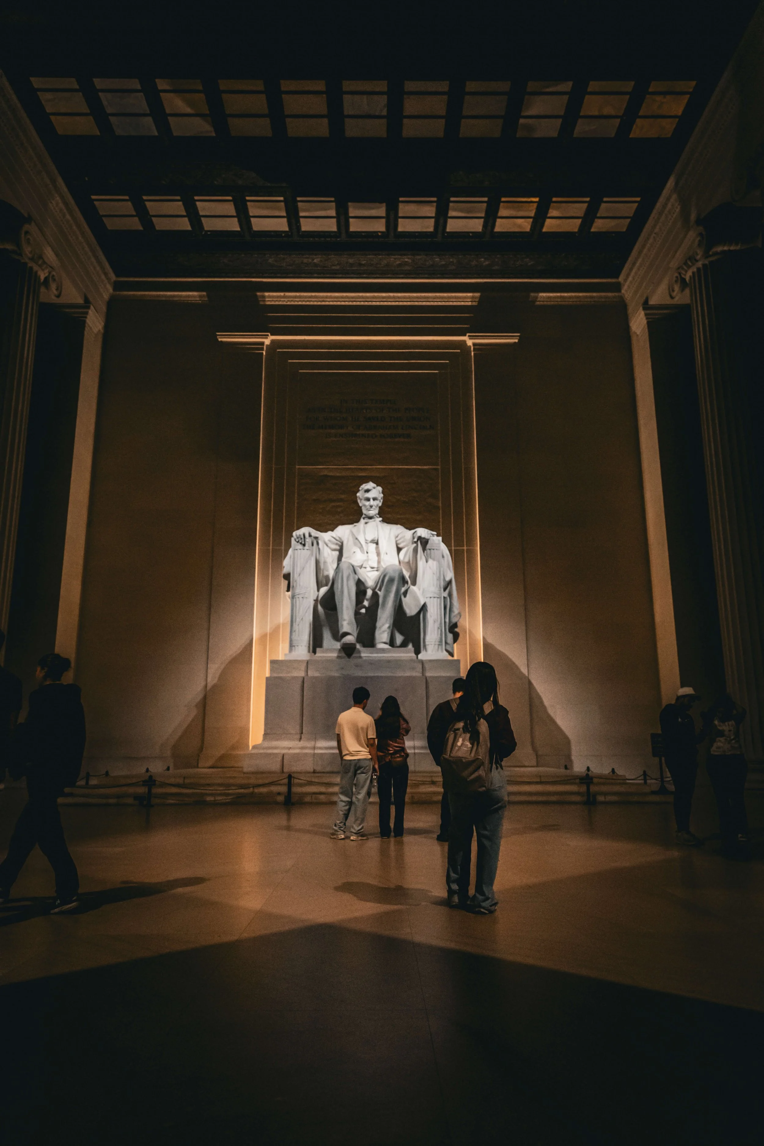 Visitors in the Lincoln Memorial in Washington, D.C., with the statue of Abraham Lincoln seated on a large chair, illuminated against the dark interior.