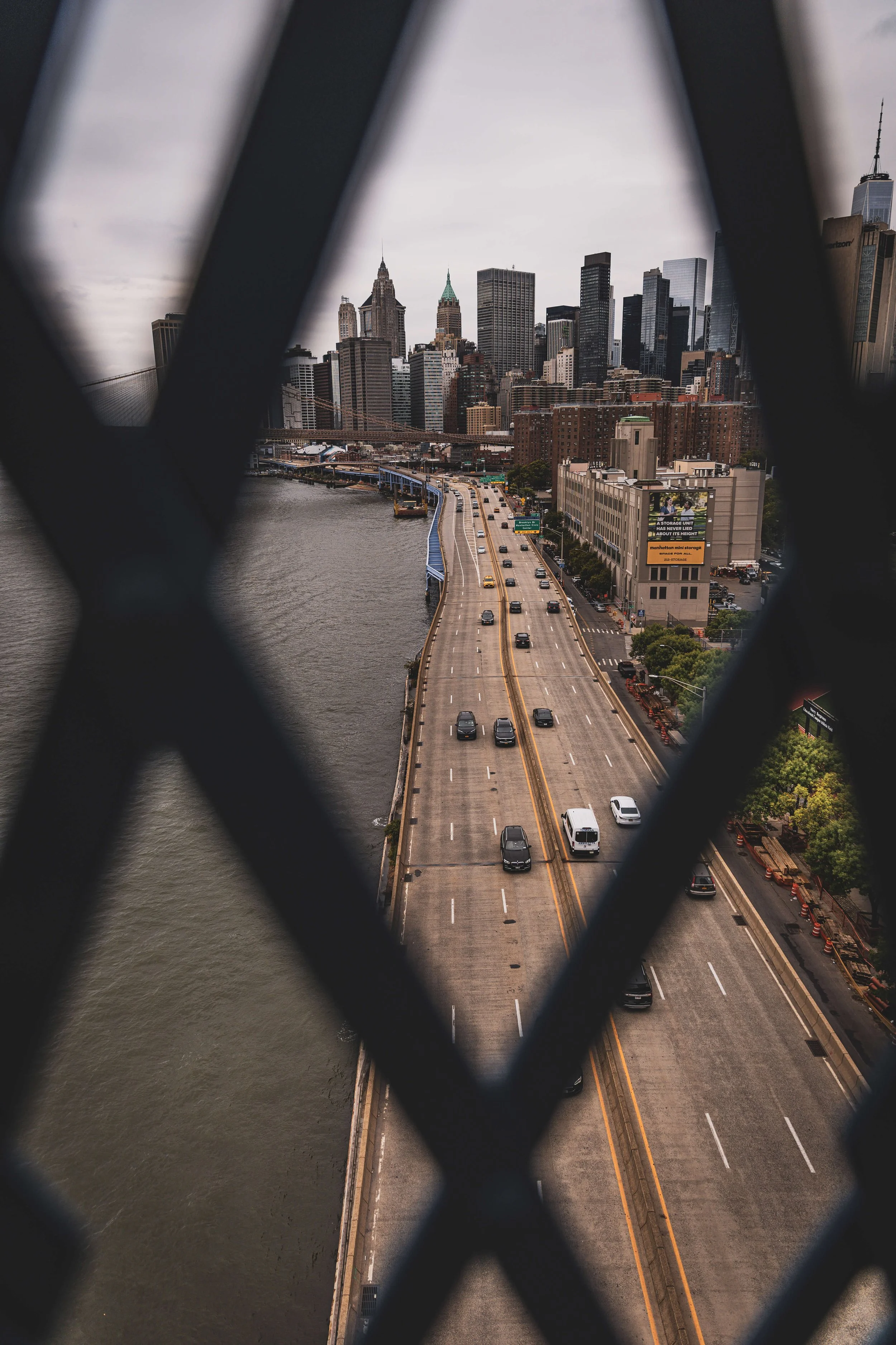 City skyline view through a metal fence, showing a river on the left and a busy highway with cars in the foreground.