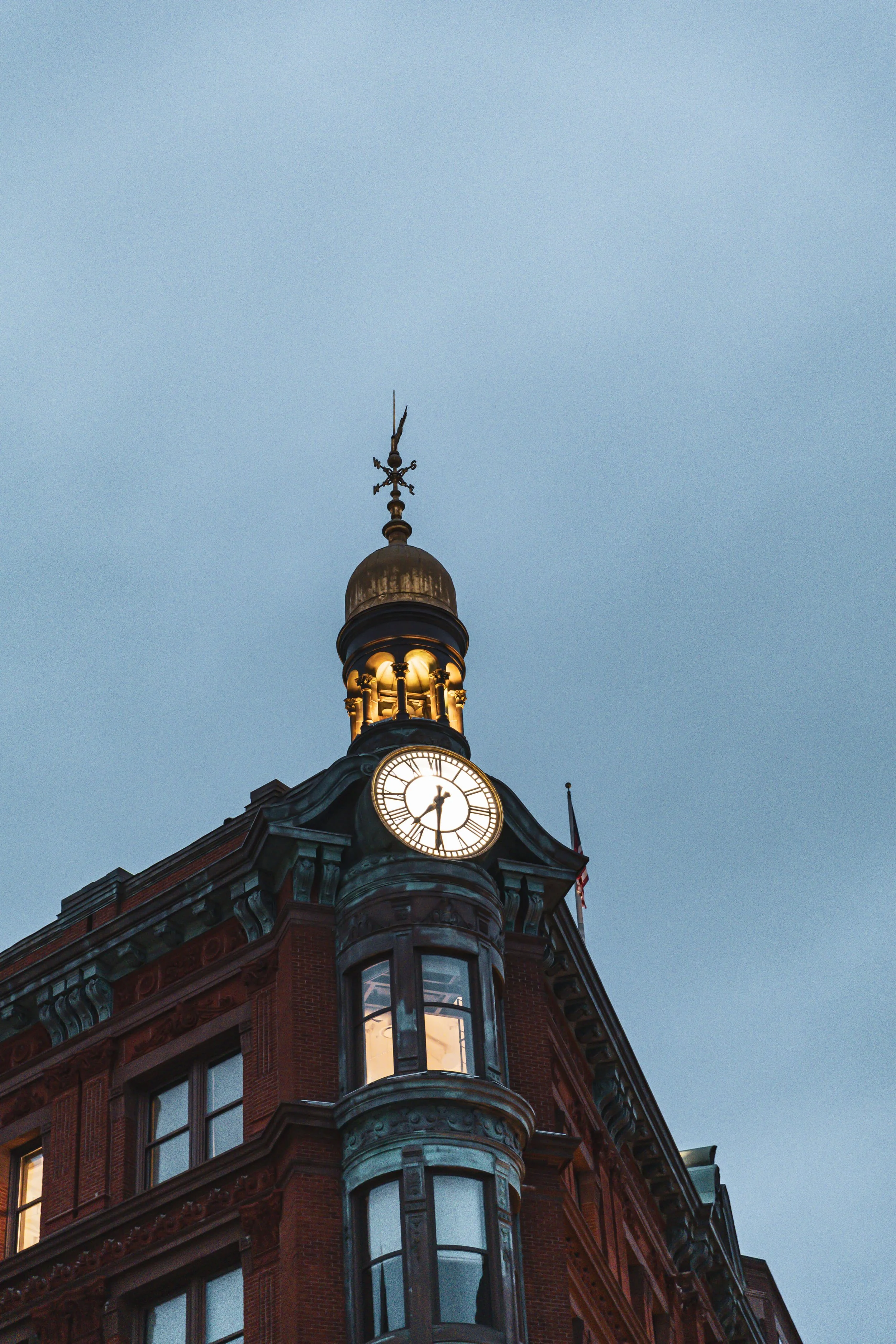 A historic brick building with a clock tower, illuminated at dusk, featuring a weather vane at the top.