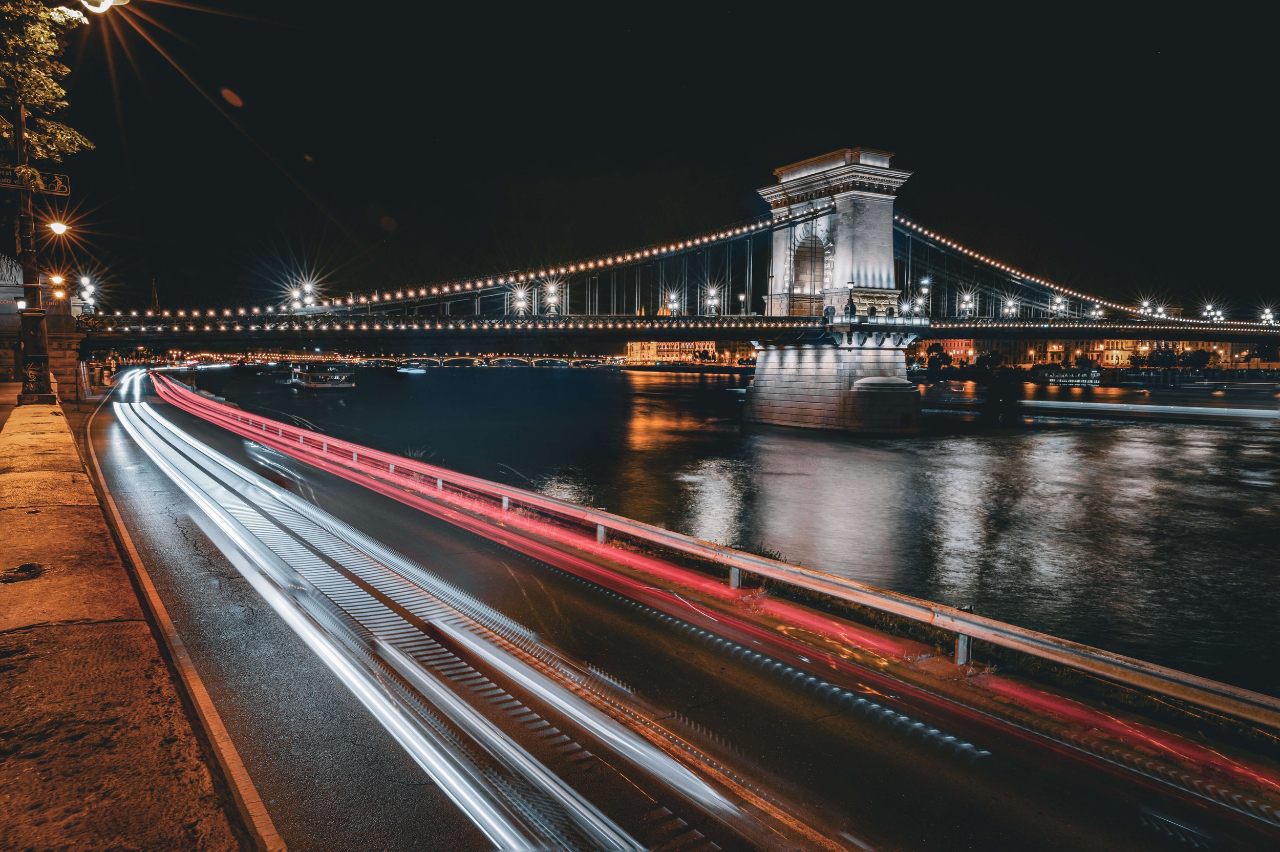 Night view of a suspension bridge illuminated by streetlights, reflecting on the river below, with light trails of moving vehicles on the road along the riverbank.